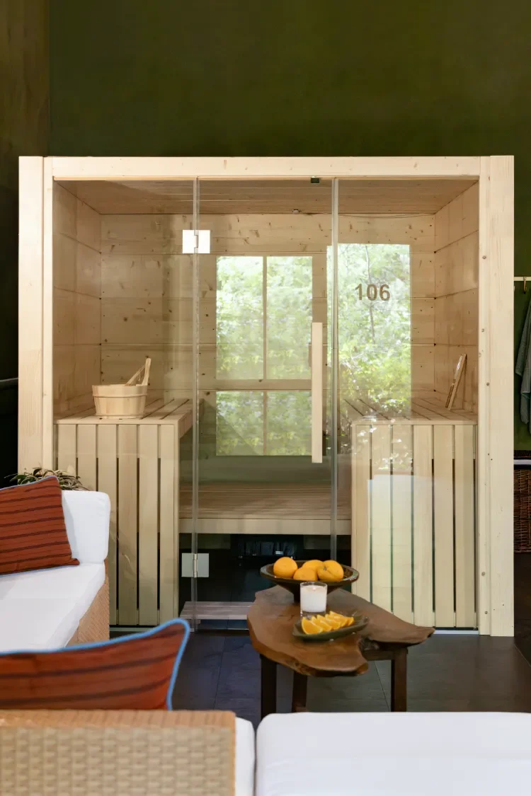 A Finnish wooden sauna room in Contrast Anerley, South London with glass doors, beside a relaxing seated area, including a wooden table with oranges and candles, and a sofa with striped cushions.