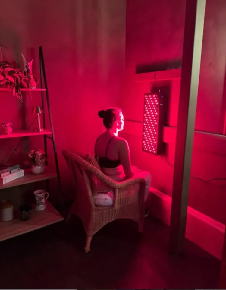 A woman in a bralette sitting in a wicker chair, facing a red light therapy device emitting red light, in a dimly lit room with wooden walls and shelves holding books, plants, and decor.