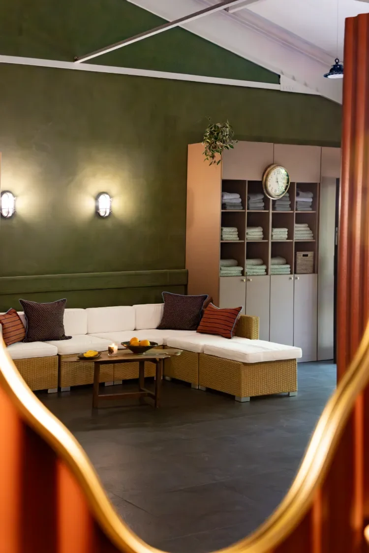 The Contrast Anerley lounge area to relax in after a sauna and/or ice bath with textured green wall, white sectional sofa, pillows, wooden coffee table with candles and fruit bowl, with folded towels, hanging plant, and soft lighting.