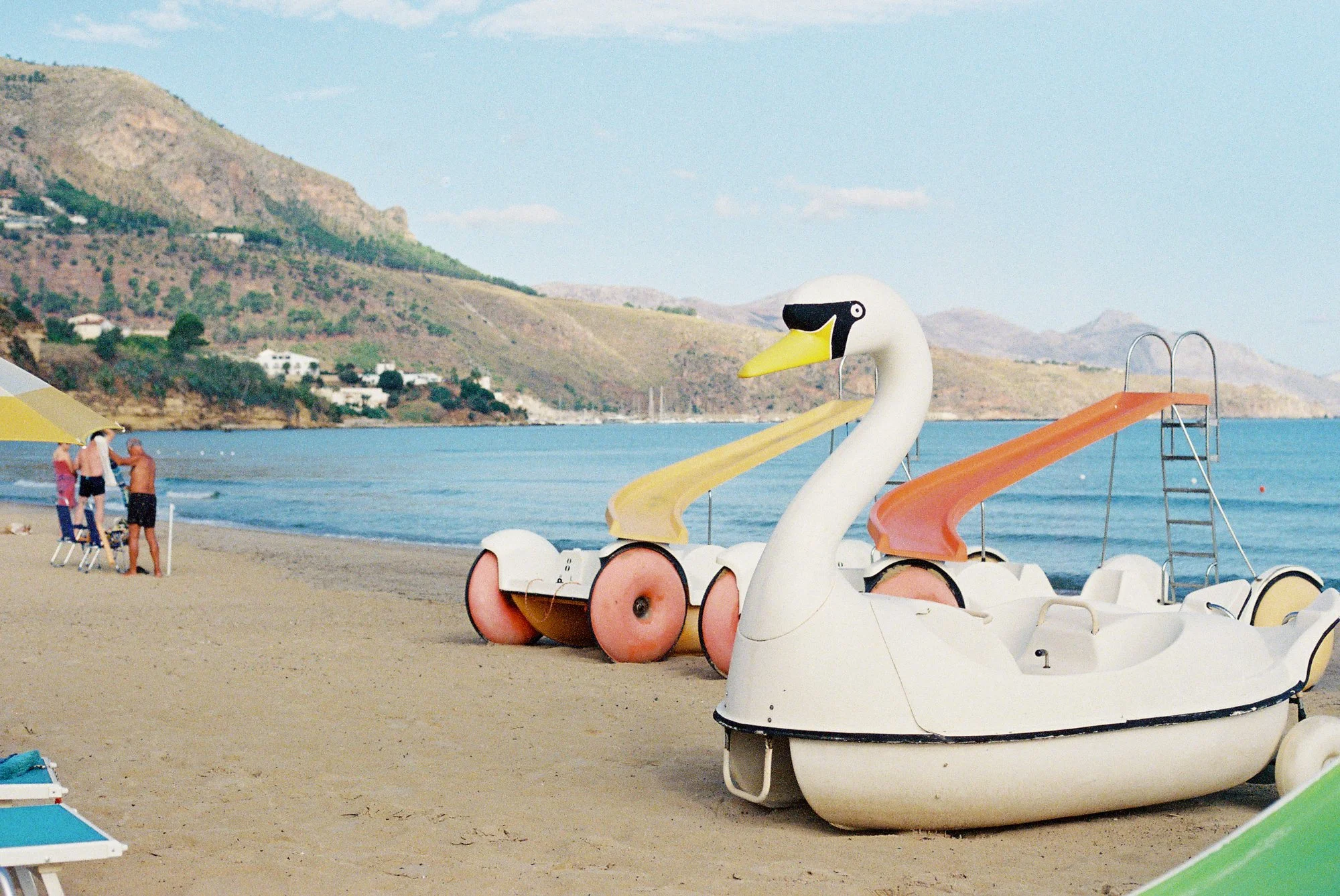Colorful swan-shaped pedal boats on a sandy beach with a yellow slide on one and an orange slide on the other, overlooking blue water and green hills under a partly cloudy sky.