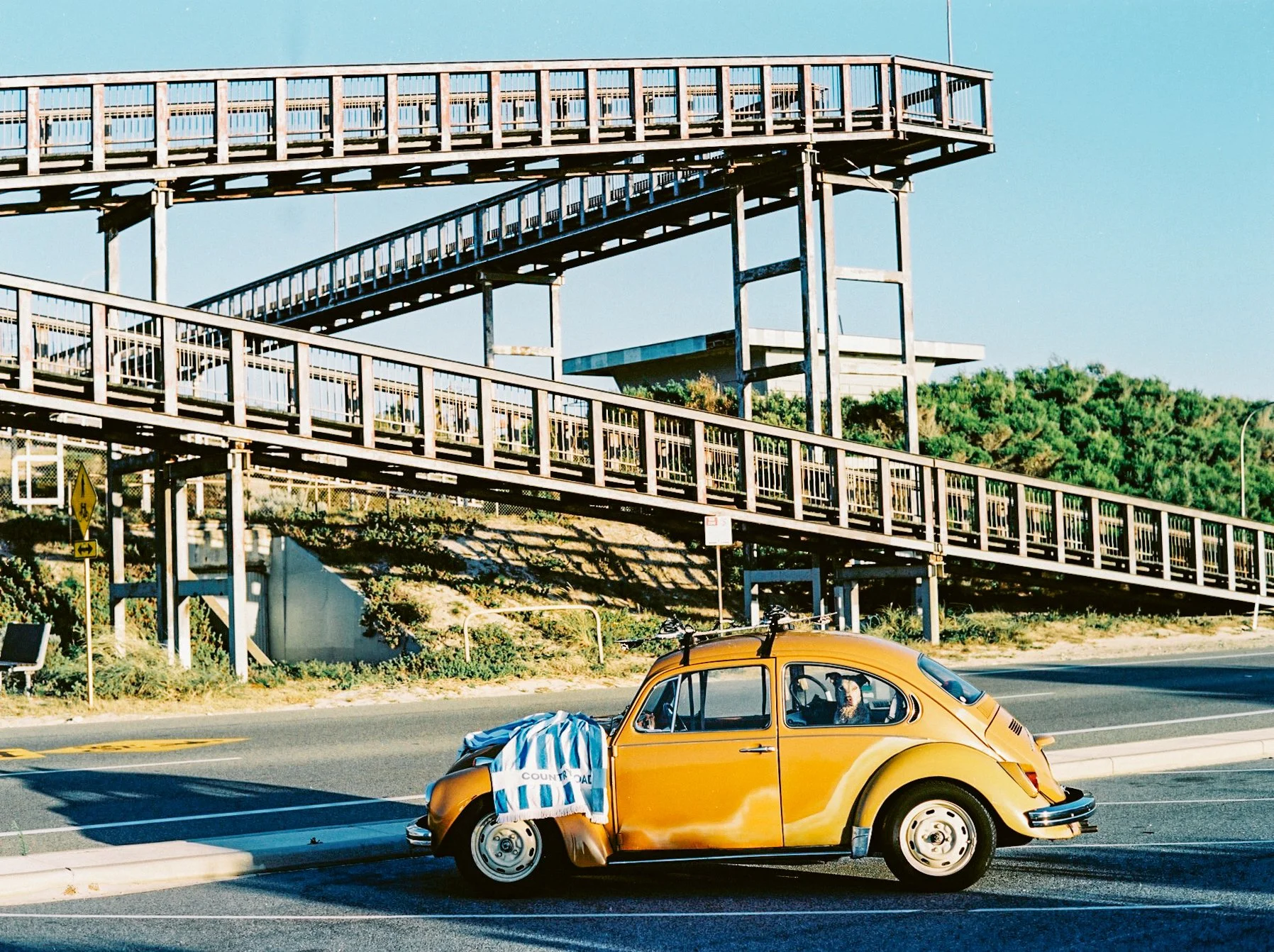 A vintage orange Volkswagen Beetle parked on the side of the road with a blue and white striped towel hanging on the front bumper. In the background, there are elevated wooden walkways and greenery.