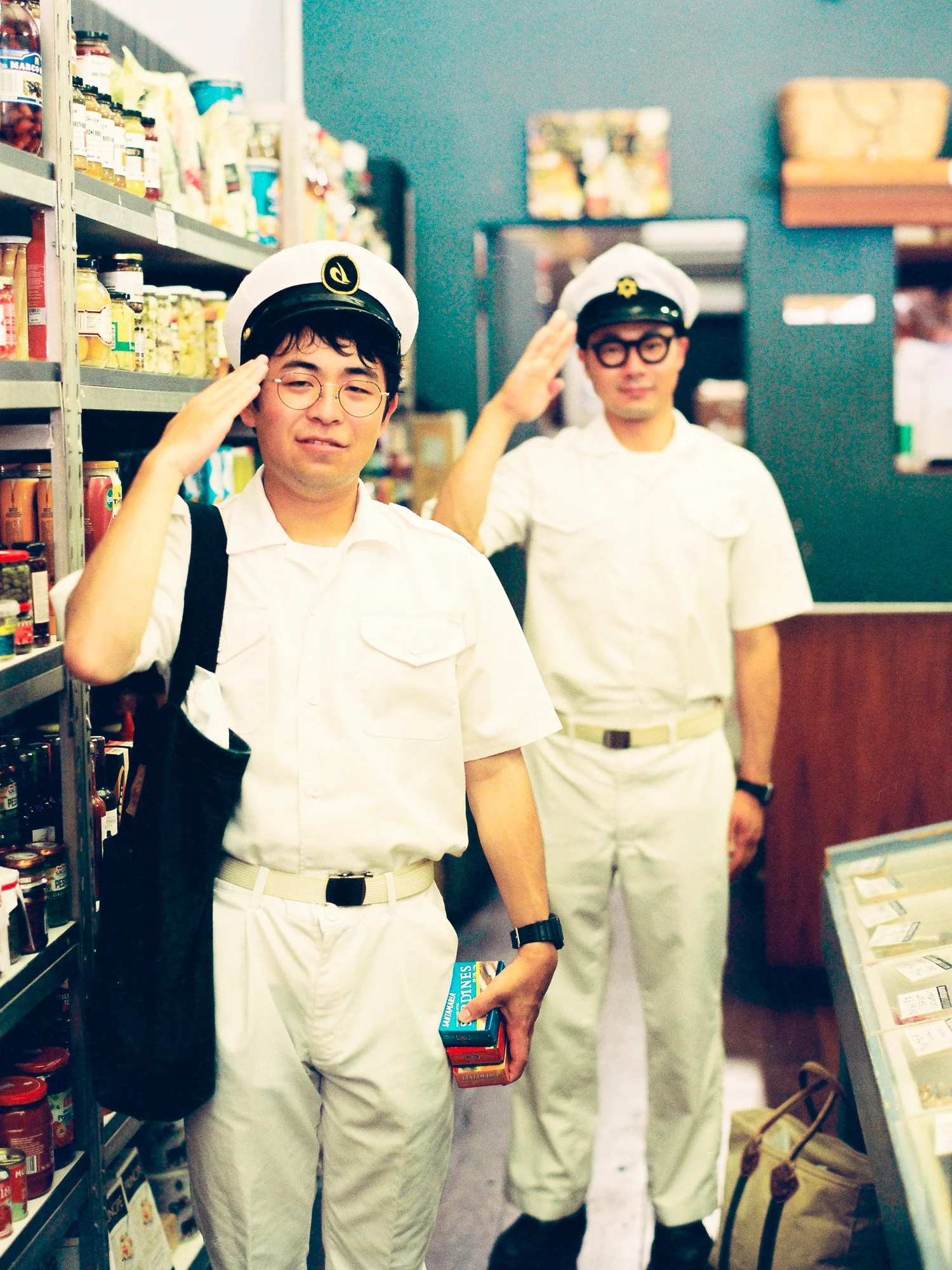 Two men dressed as sailors in white uniforms with hats, saluting inside a grocery store aisle.