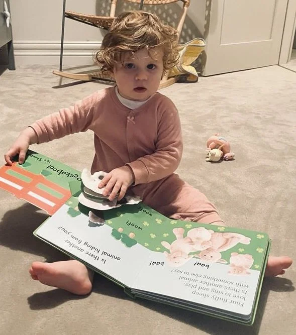 Hatty, a child with curly hair and a pink outfit sitting on a carpeted floor, holding a children's book open with colourful illustrations of bunnies and a fox.