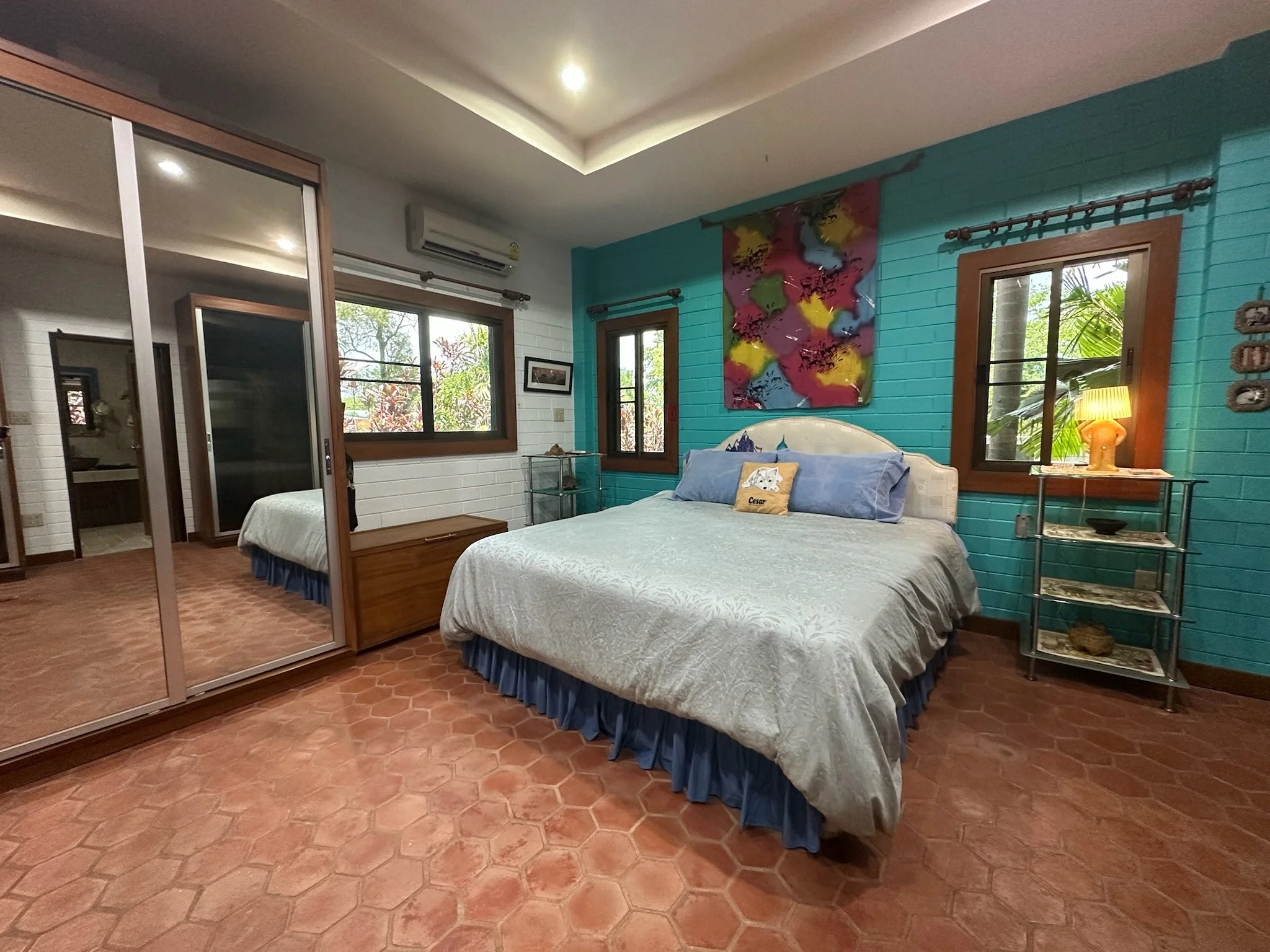 A bedroom with a blue accent wall, a wooden bed with red and colorful bedding, framed artwork, a window with white curtains, a small dining table with chairs, and a mirror reflecting part of the room.
