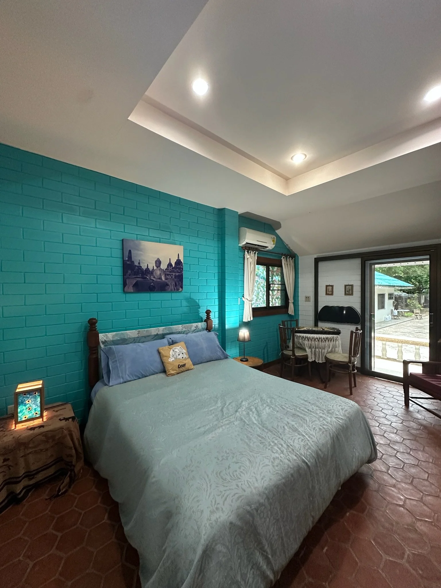 A brightly decorated bedroom with blue brick walls, a white bed with black and white patterned blanket, colorful wall art, red curtains, and a window