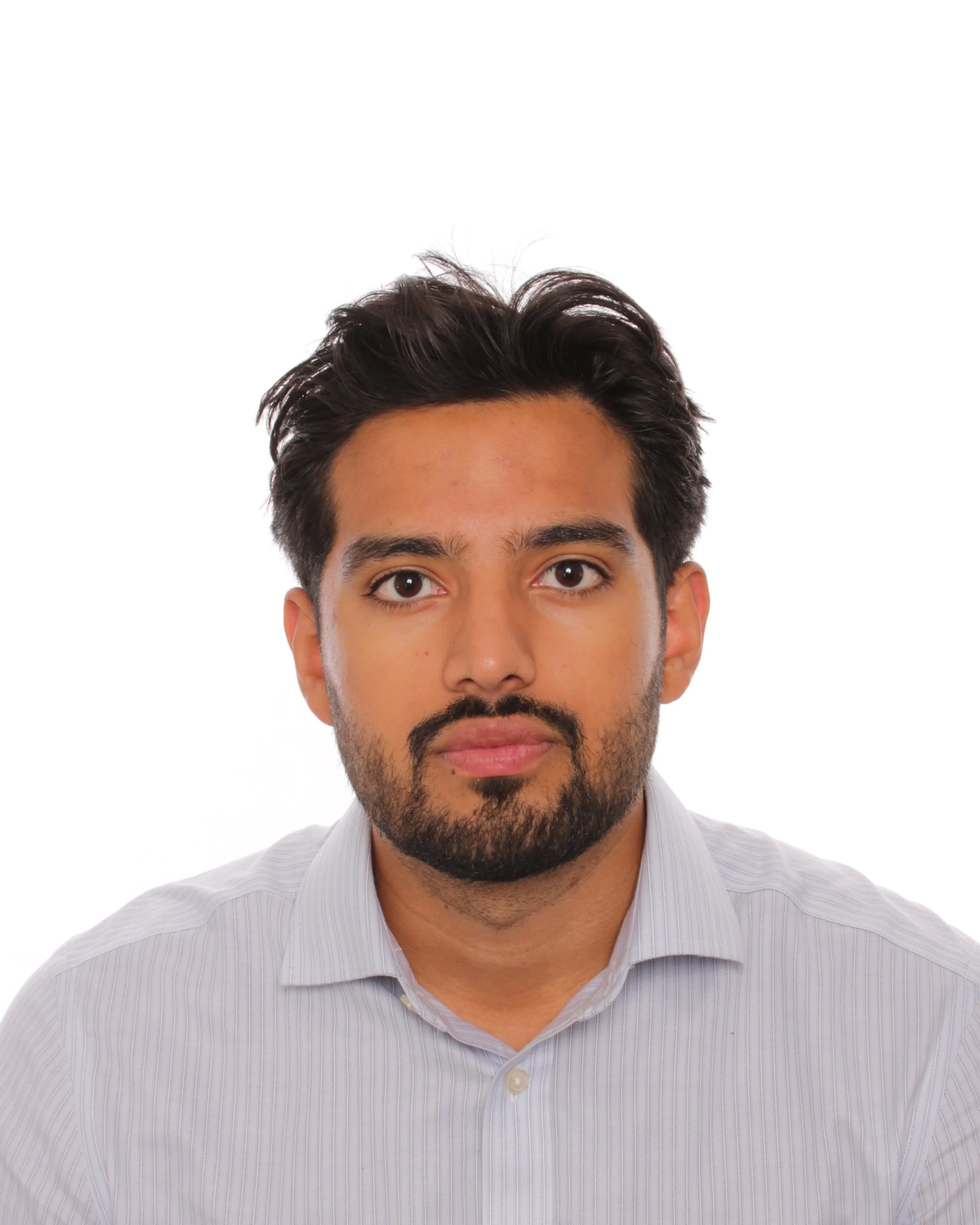 A young man with short black hair, a beard, and a mustache, smiling in front of a beige wall with a dark brown border and some hanging wires.
