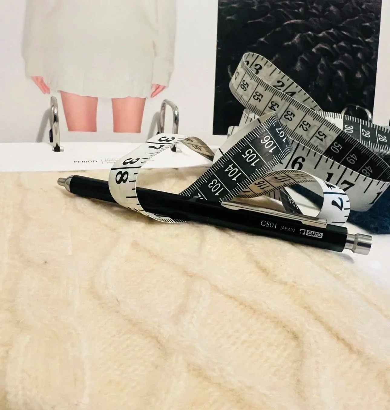 A black pen, a measuring tape, and a photograph of a person with nude legs wearing a cream-colored coat are on a light-colored surface, with the measuring tape partly looped around the pen and the photo.