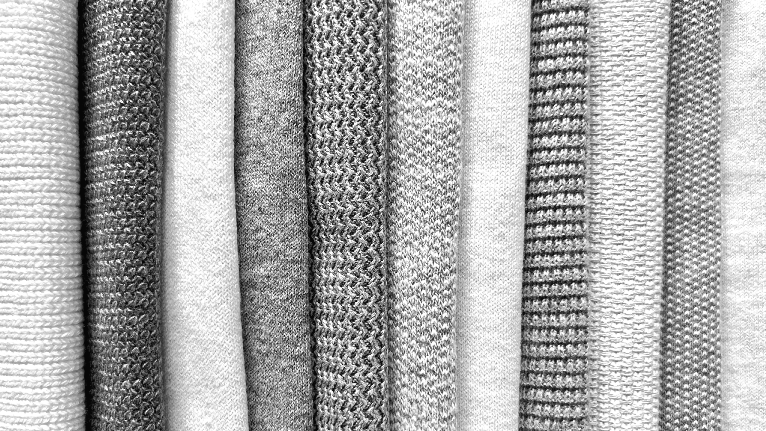 A collection of various knitted swatches folded vertically, featuring different textures and patterns in shades of gray. 14-9 gauge knitwear.
