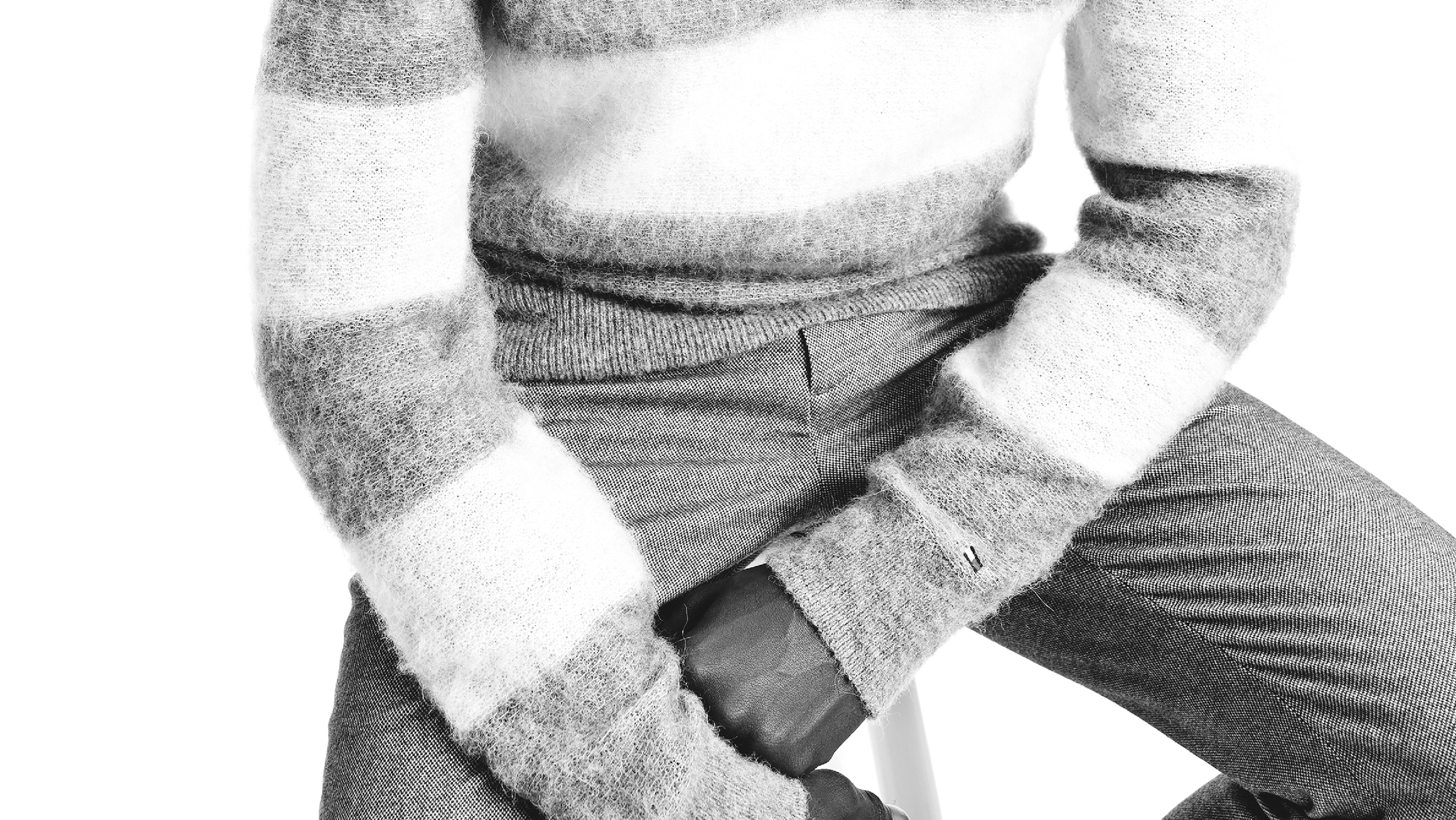 A person wearing a striped sweater and patterned pants sitting with hands clasped over a small bag on their lap, in black and white.