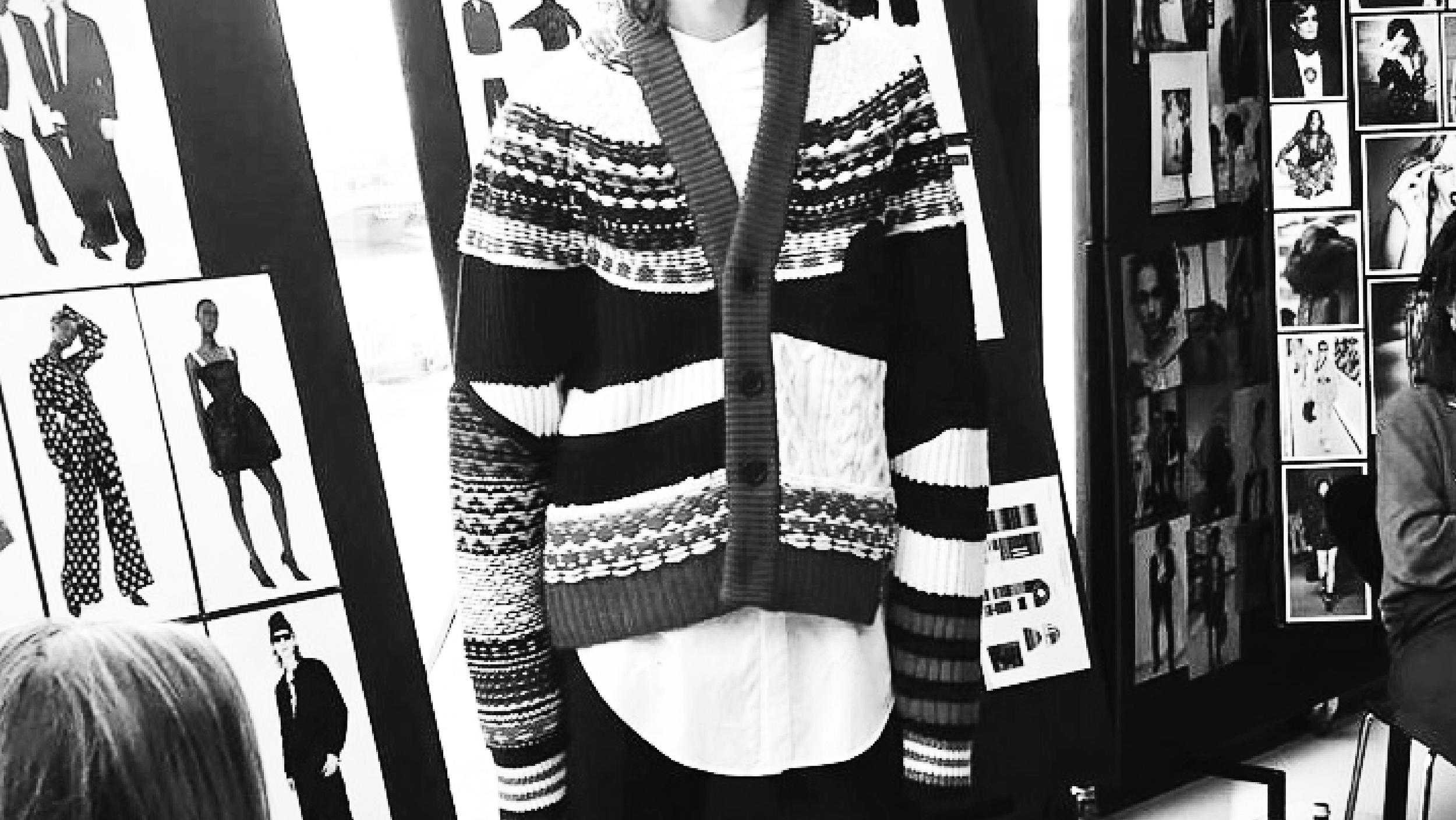Black and white photo of a woman in a patterned sweater, standing in front of a collage of fashion sketches and photographs displayed on boards.