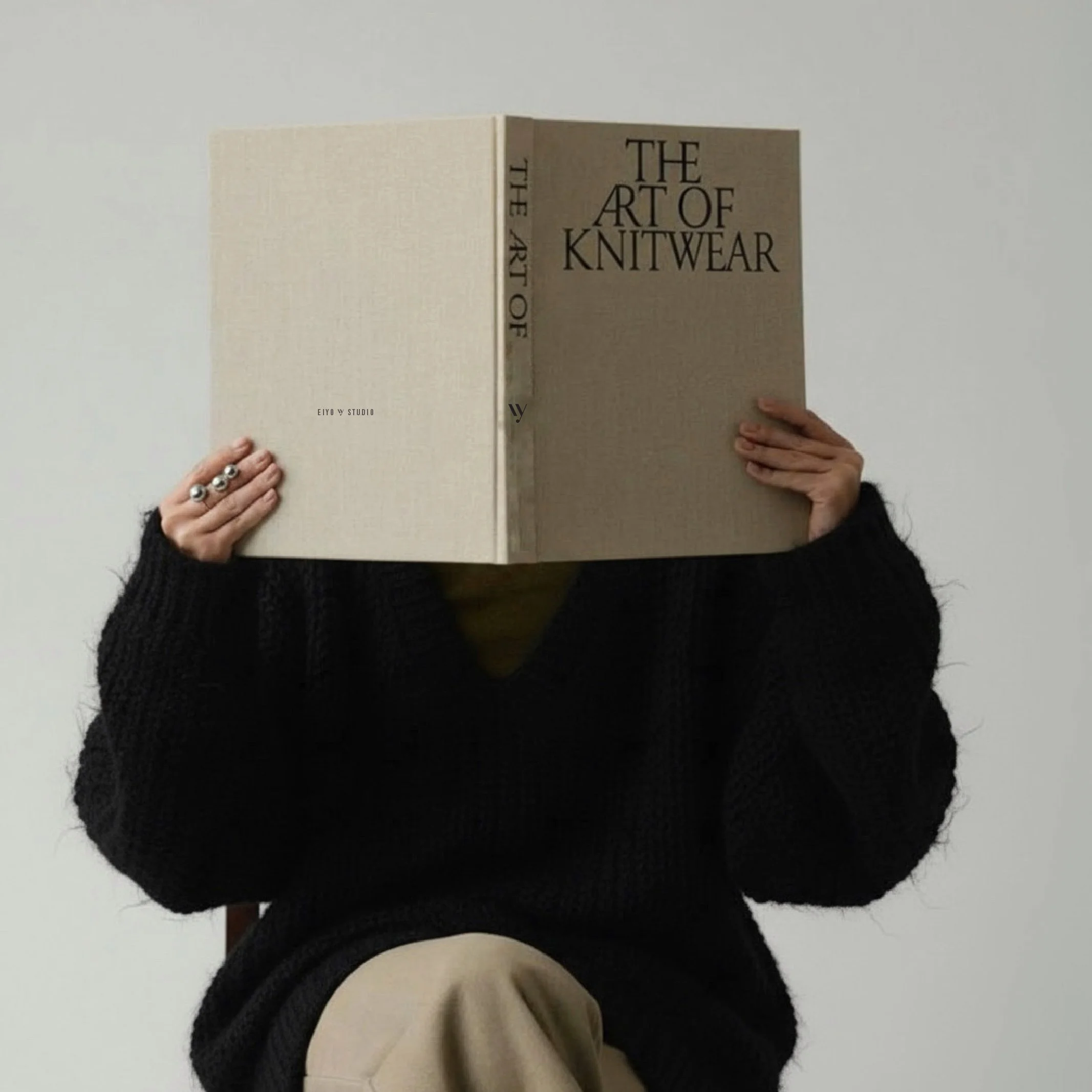 Person sitting in front of a plain white wall, holding an open book titled 'The Art of Knitwear' in front of their face. The individual is wearing a black sweater and beige pants, with rings on their left hand.