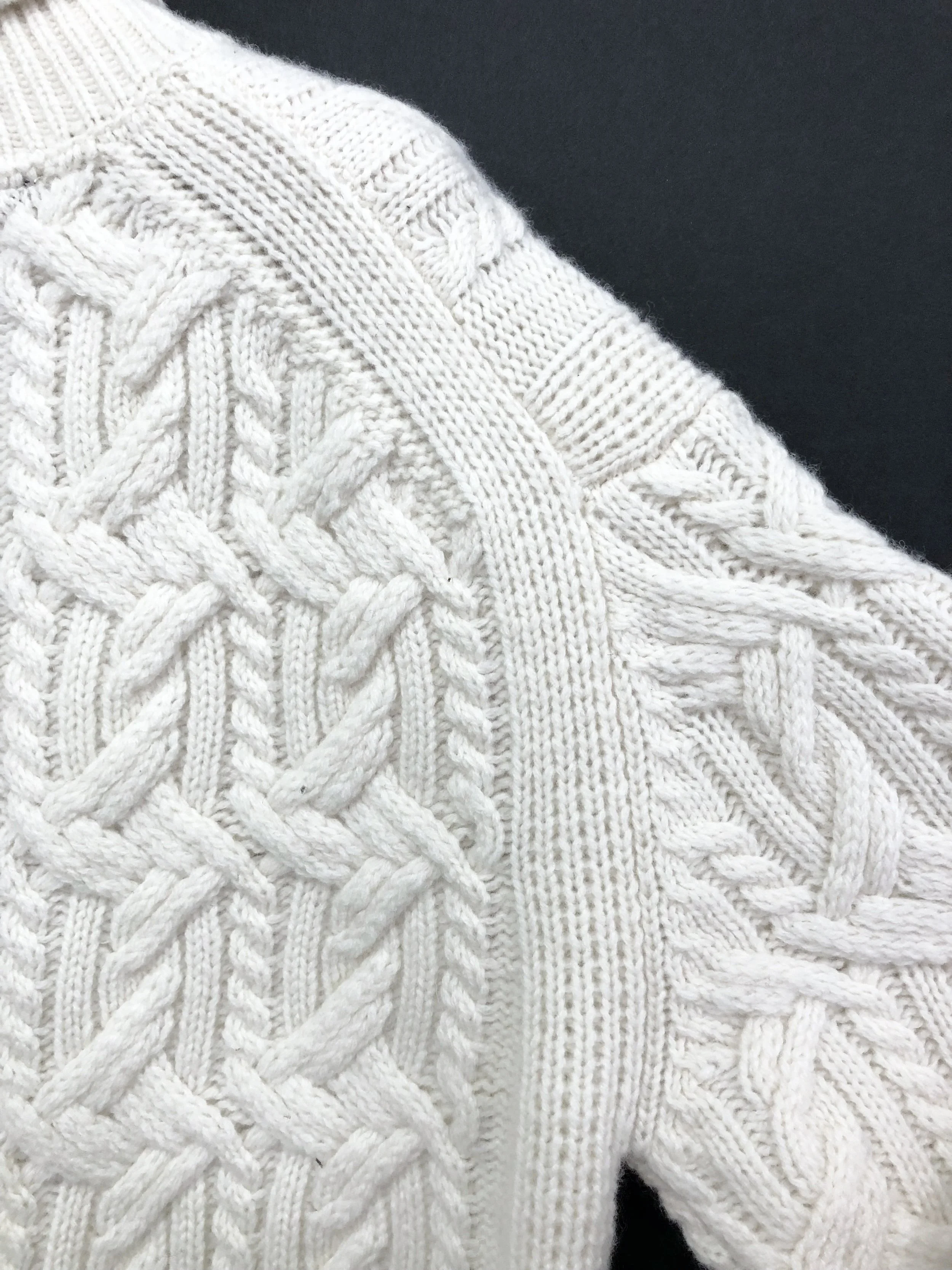 Close-up of a white cable-knit sweater with intricate patterns, including braids and textured stitches.
