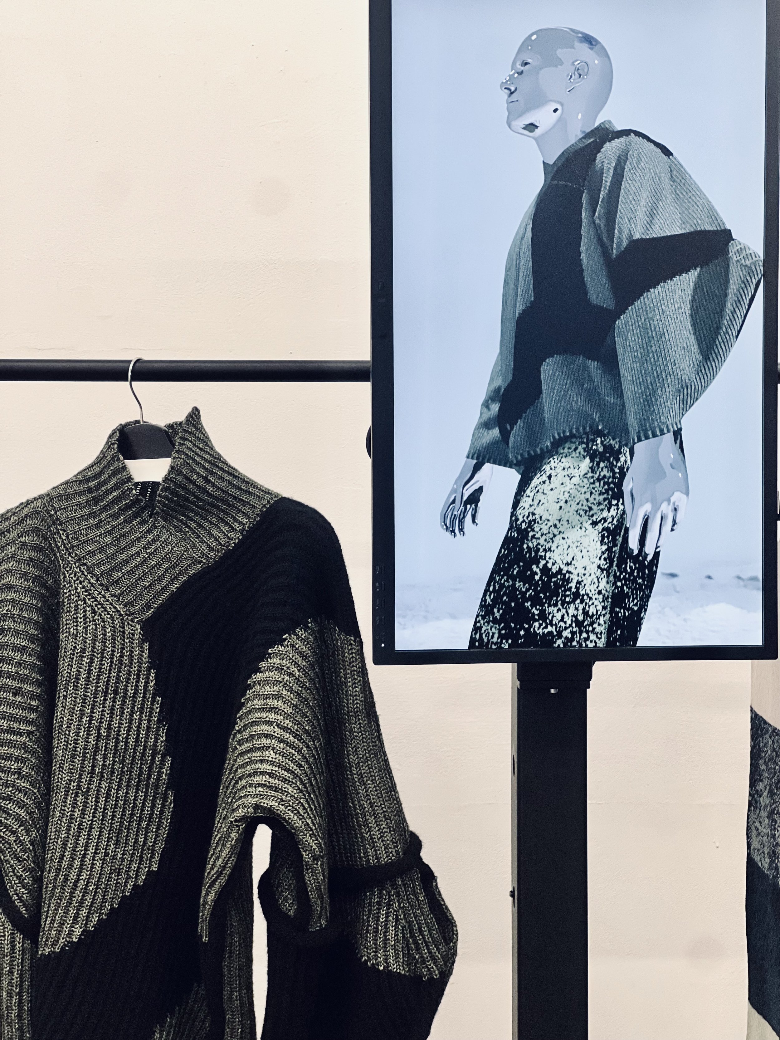 Display of a black and gray knit sweater on a hanger with a digital screen showing a model wearing a gray olive and black patterned sweater. full look 3D simulation made with CLO3D by KnitwearLab.