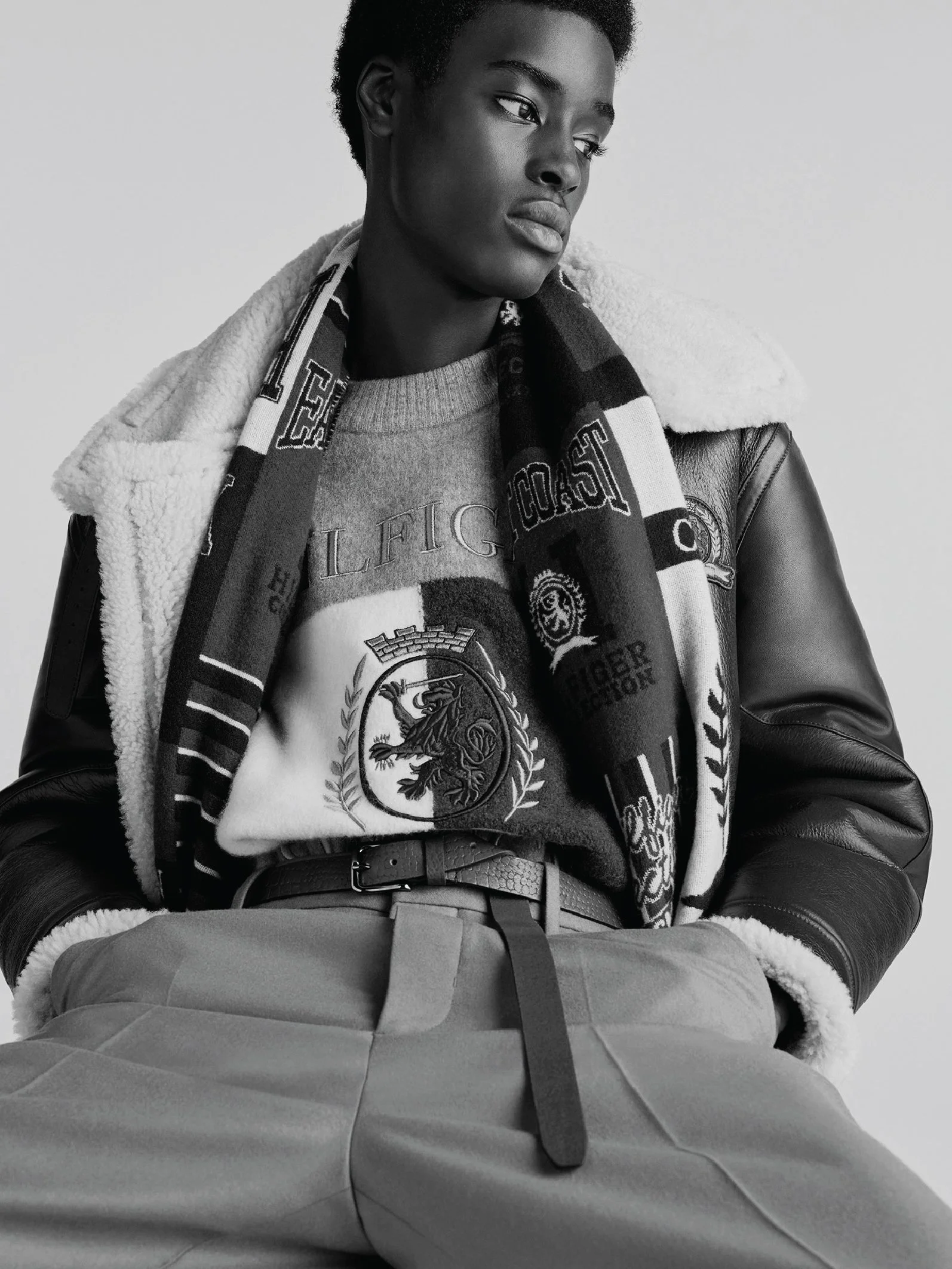 Black and white portrait of a young man with short curly hair, wearing a graphic sweater, a scarf, a shearling-lined jacket, and high-waisted pants with a belt, seated with hands in pockets and looking to the side.