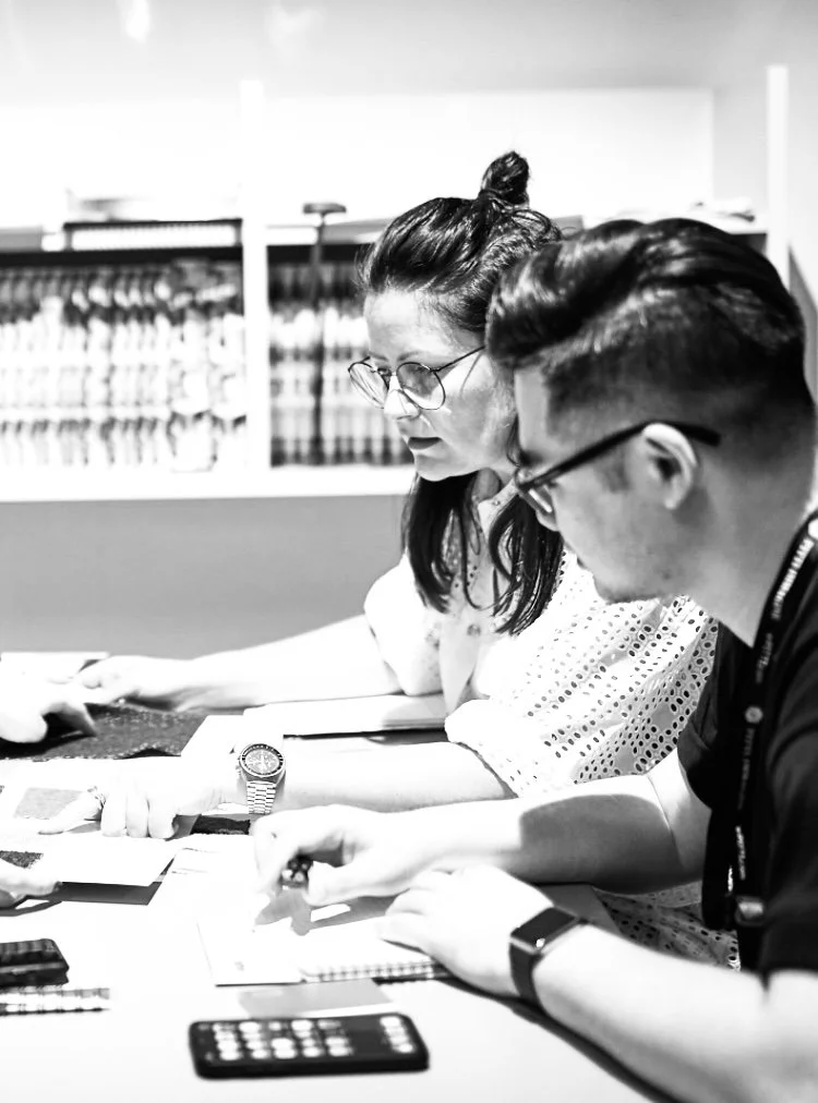 Two people, a woman with glasses and a man with glasses, sitting at a table looking at documents or a phone in a library or office setting.