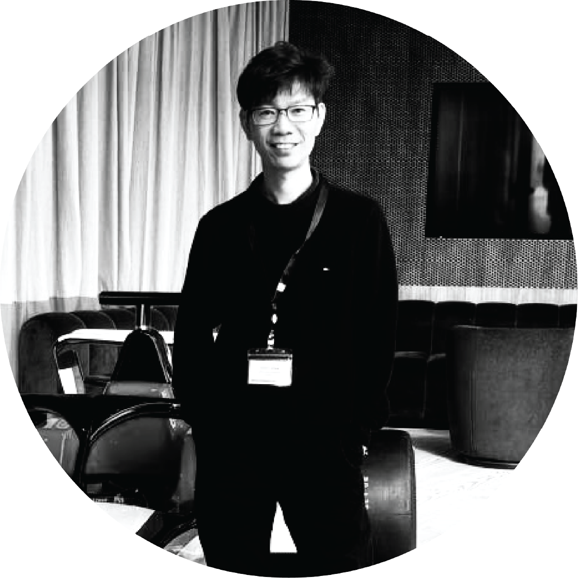 A young man with glasses and dark hair, smiling, standing in a room with curtains, chairs, and a TV in the background.