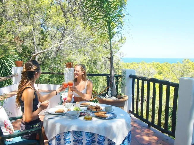 Two women enjoying a meal on a terrace in the retreat centre in Ibiza with a view of trees and the ocean, surrounded by lush greenery and bright sunlight, sharing a toast with drinks.