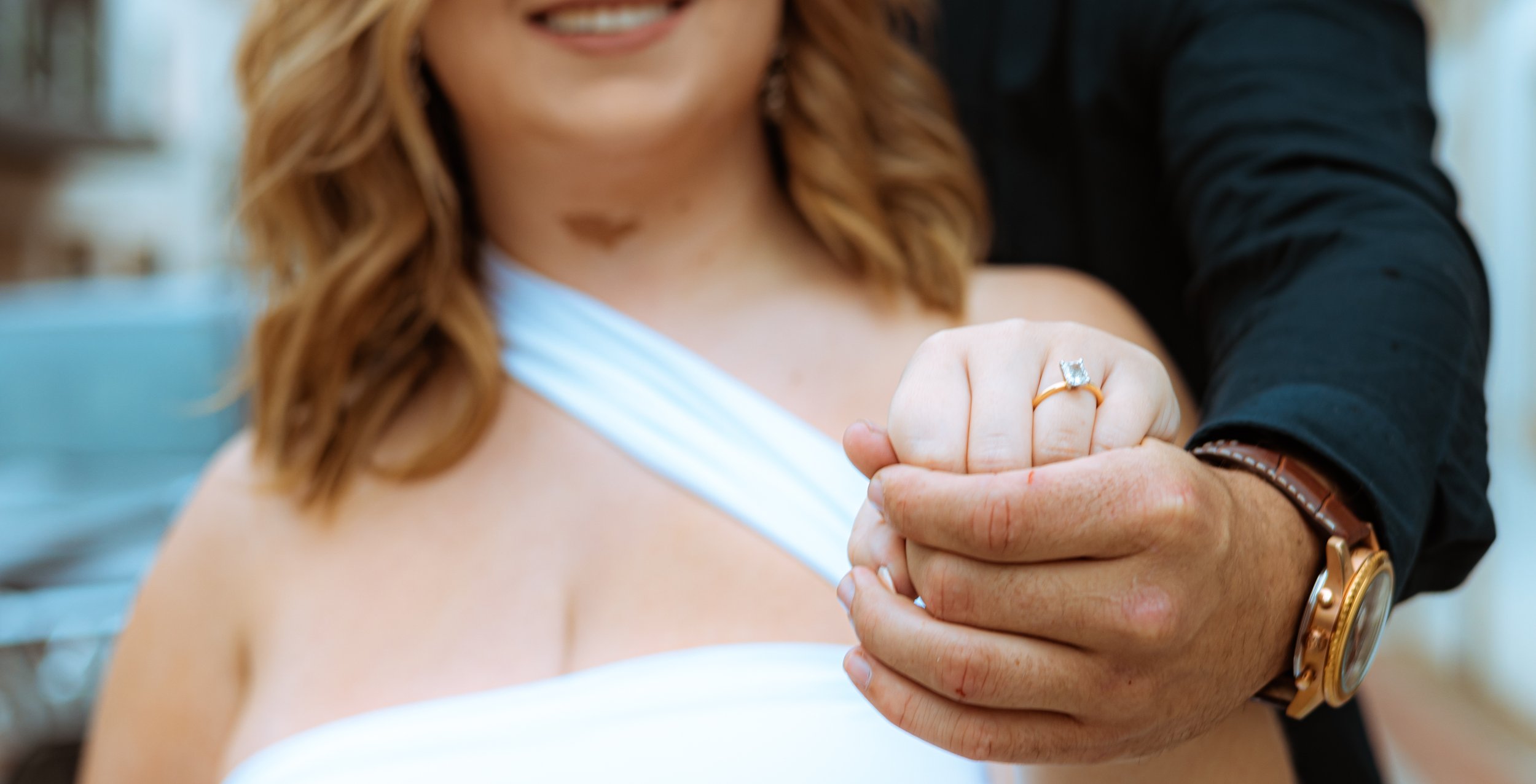 Close-up of a woman wearing a white dress and an engagement ring on her finger, being held by a man dressed in black. The woman is smiling, and the scene suggests a romantic or engagement moment.