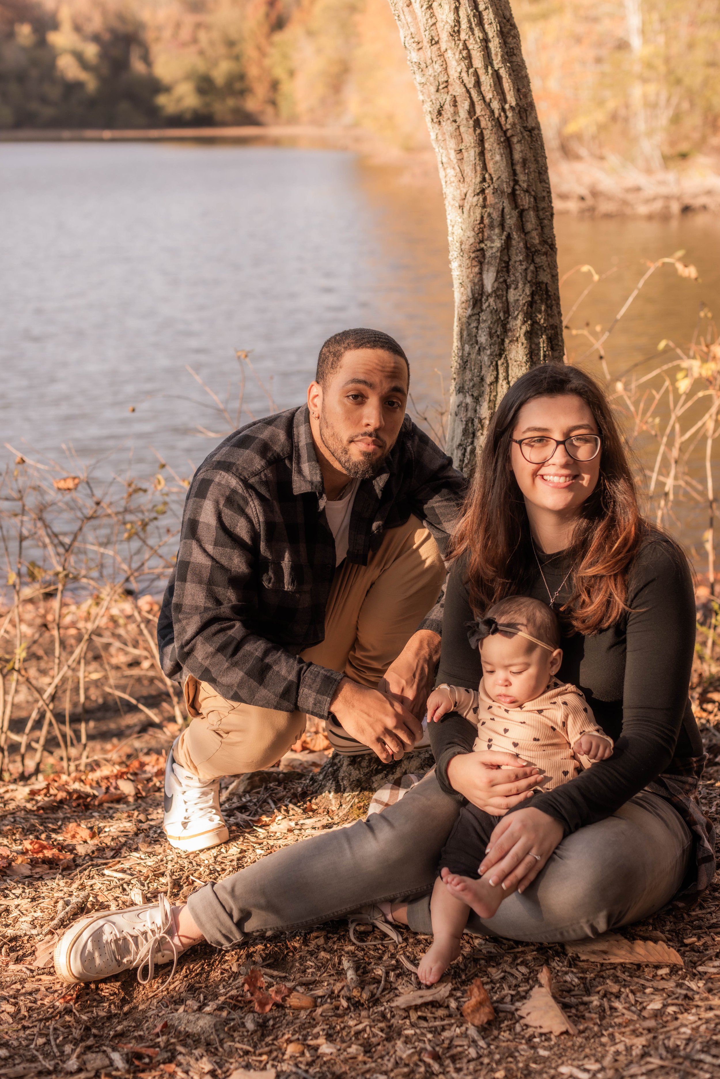A family of three sitting on the ground near a lake with autumn foliage in the background, including a man, a woman, and a young girl, all smiling and enjoying a fall day outdoors.
