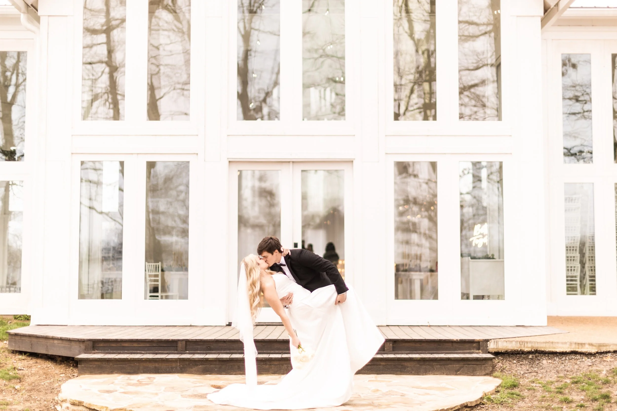 A bride and groom sharing a kiss on a small stone patio outside a white building with large glass windows.