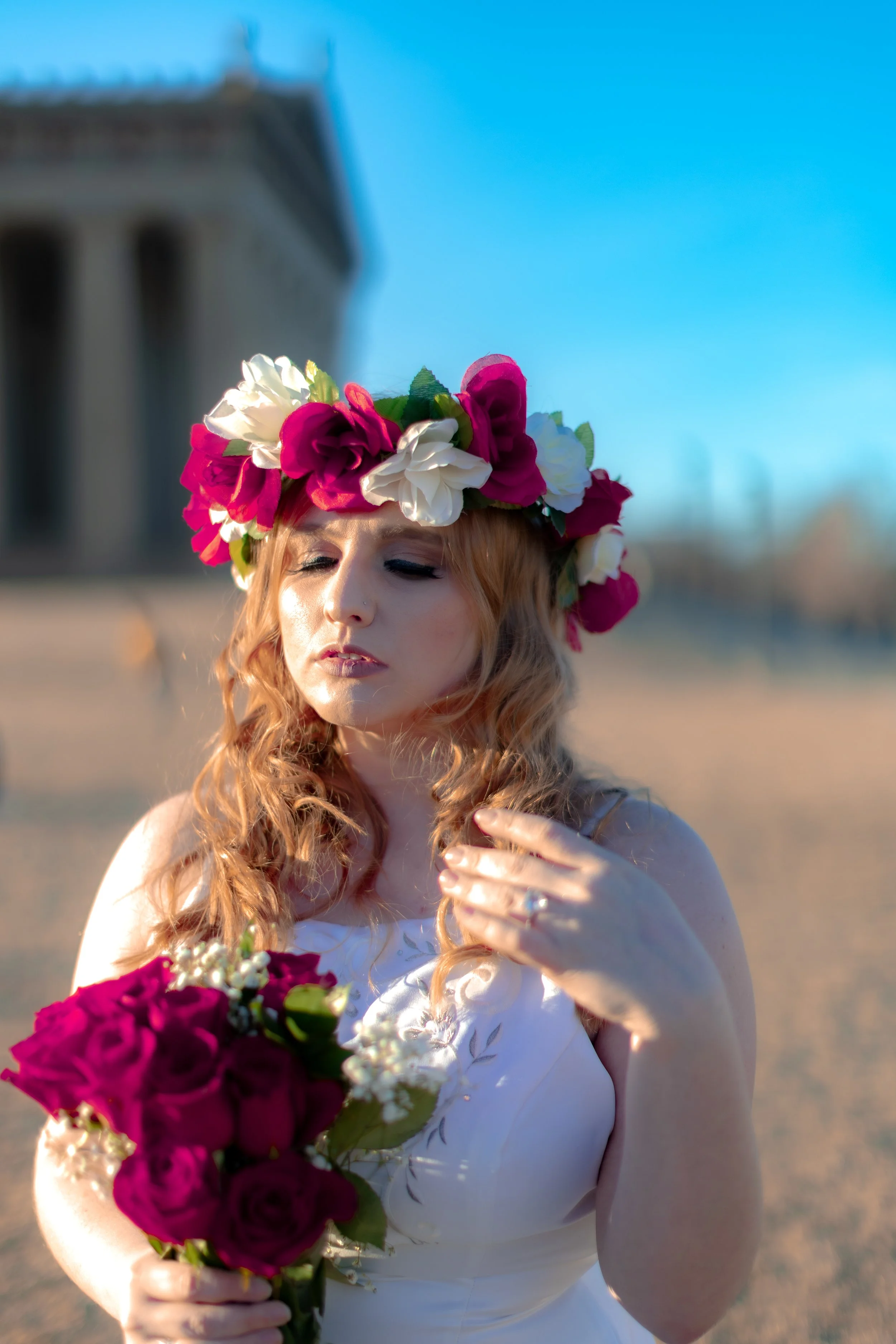 A woman with curly red hair wearing a floral crown made of pink and white flowers, holding a bouquet of pink roses, standing outdoors with a blurred building and blue sky in the background.