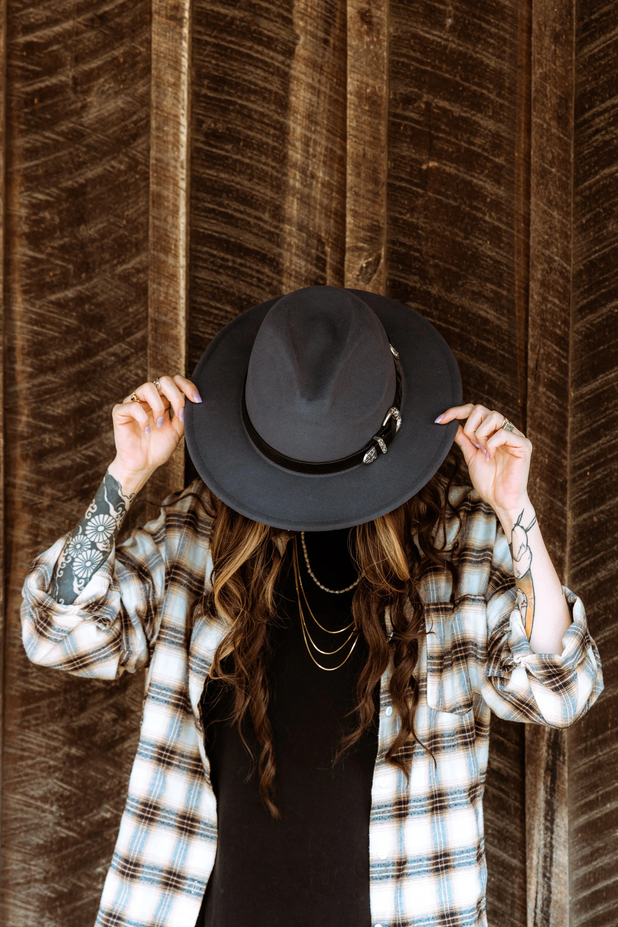 Woman with long wavy hair wearing a plaid shirt and multiple gold necklaces, holding a black wide-brimmed hat, standing against a wooden wall.