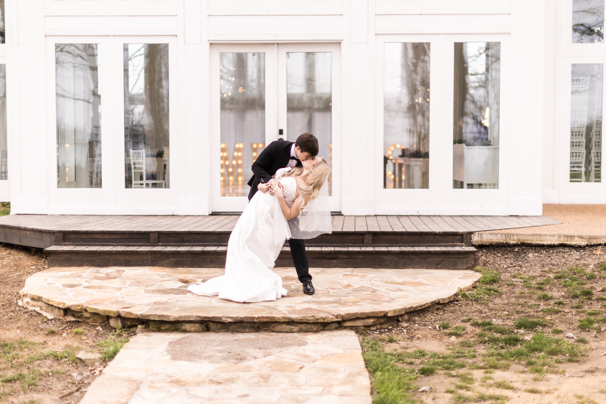 A bride and groom in wedding attire sharing a romantic moment outside a white building, with the groom dipping the bride and kissing her.