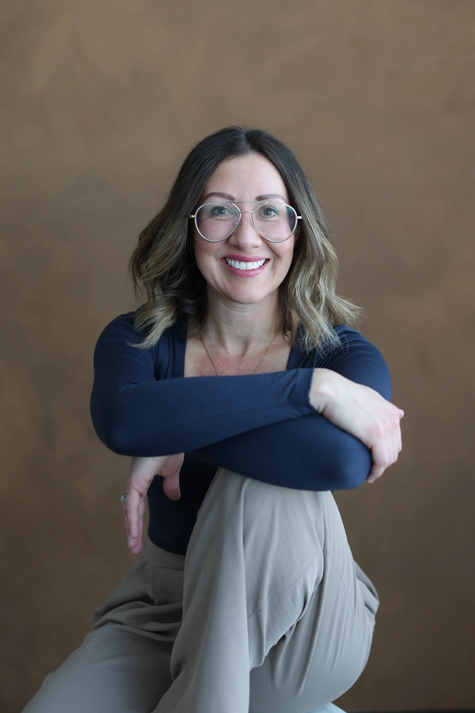 A woman with shoulder-length wavy hair, glasses, and a bright smile sitting in front of a plain brown background. She is wearing a navy blue long-sleeve top and beige pants, with her arms crossed and resting on her knees.