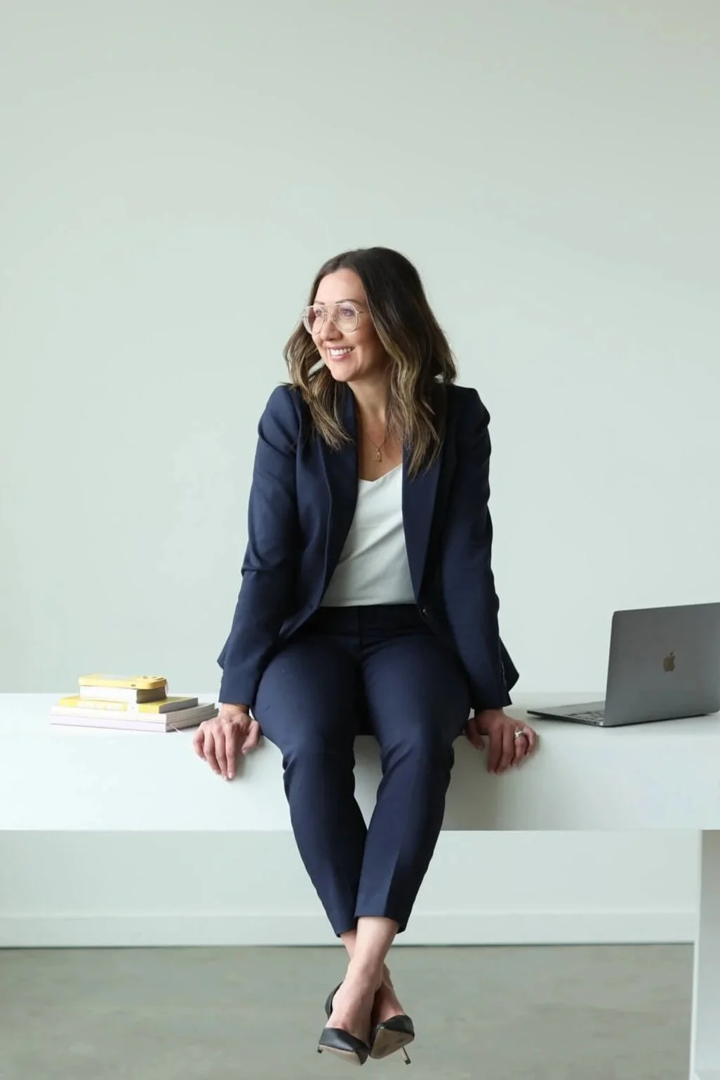 Woman in a navy blue suit sitting on a white desk with a laptop, stacks of books, and yellow sticky notes, smiling and looking to her right against a plain light-colored wall.
