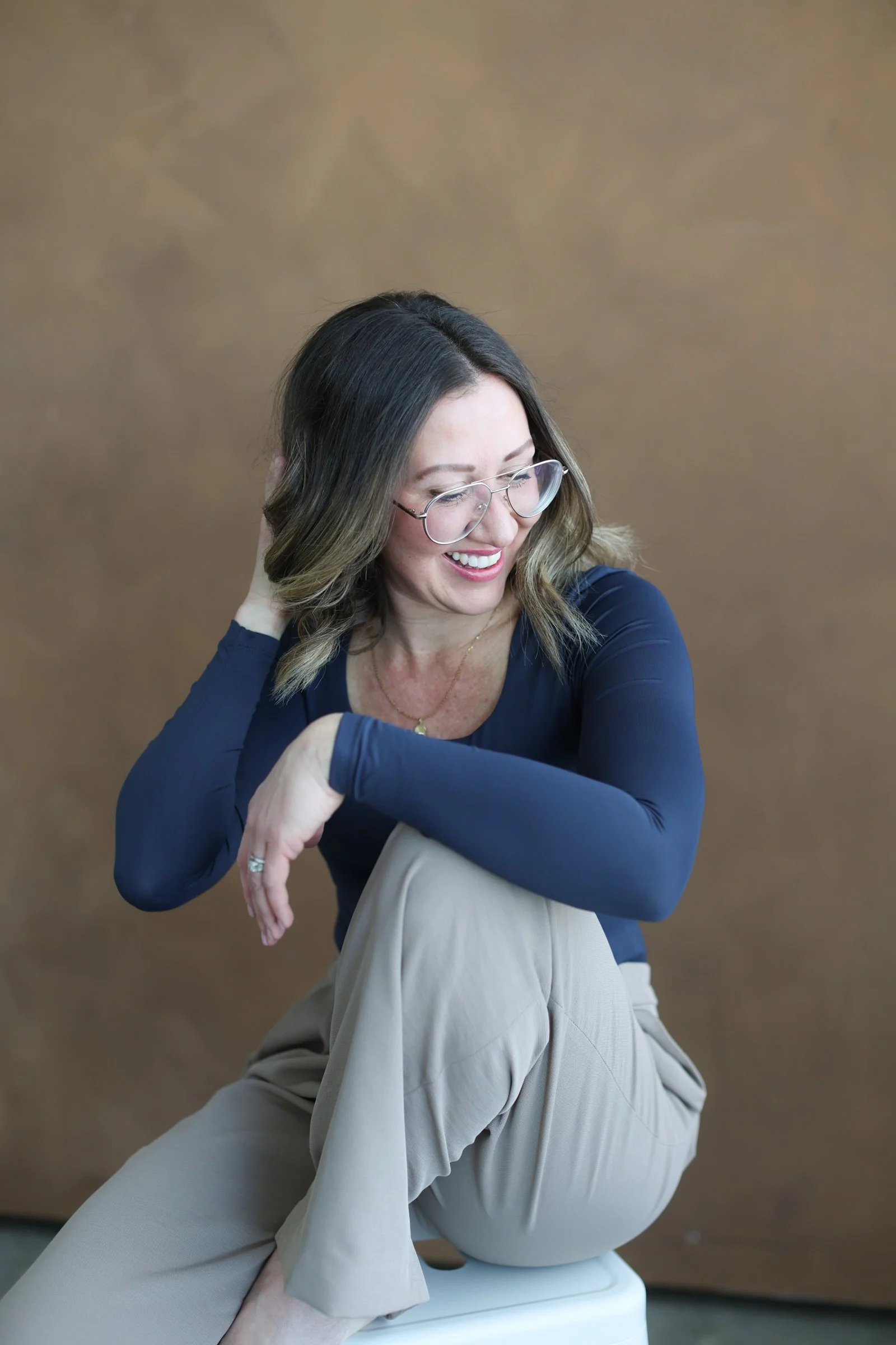 A woman with shoulder-length brown hair, wearing glasses, a navy blue long-sleeve shirt, and beige pants, is sitting on a white toilet with her leg propped up on the seat. She is smiling and looking down, with her right hand touching her hair, in a room with a brown background.