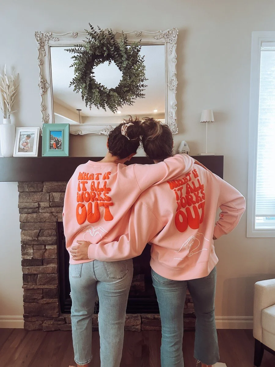 Two women in jeans standing with backs to the camera wearing matching pink jumpers that say "what if it all works out?" in front of a fireplace.