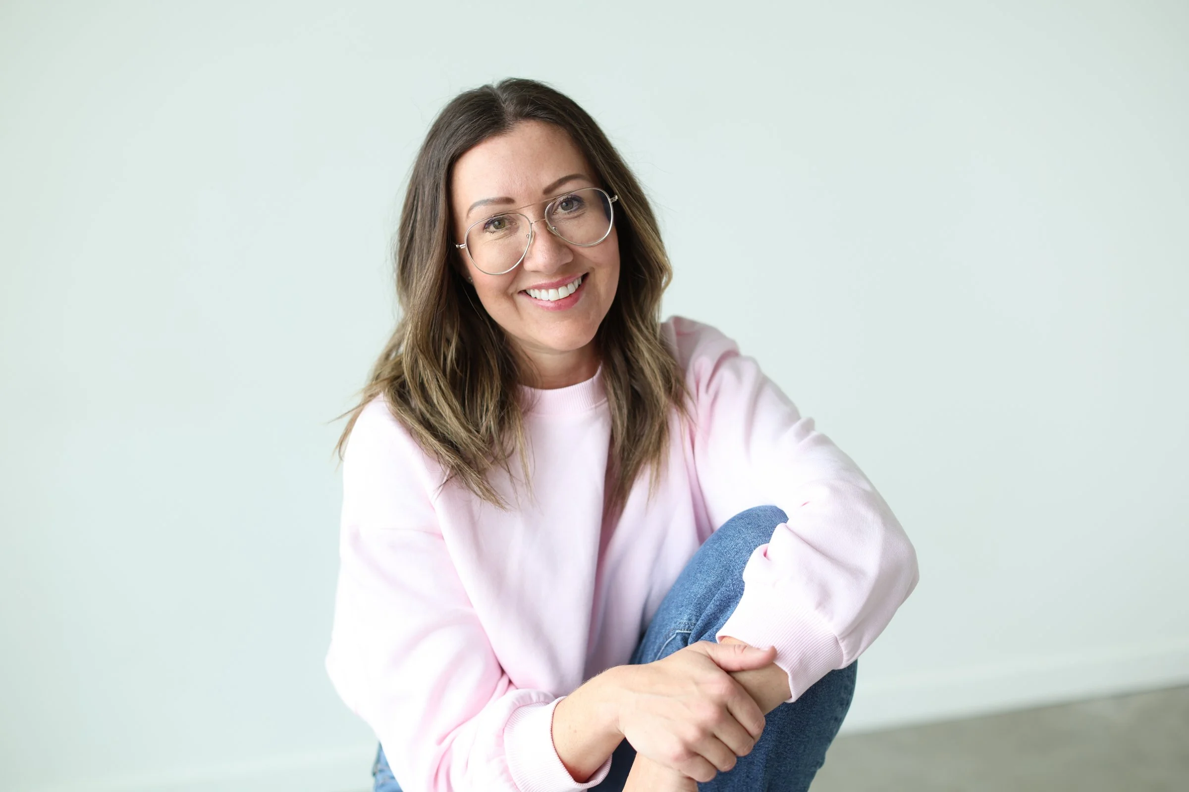 A young woman with brown hair wearing glasses and a pink sweatshirt, sitting with her knees drawn up, smiling and looking at the camera against a light green background.
