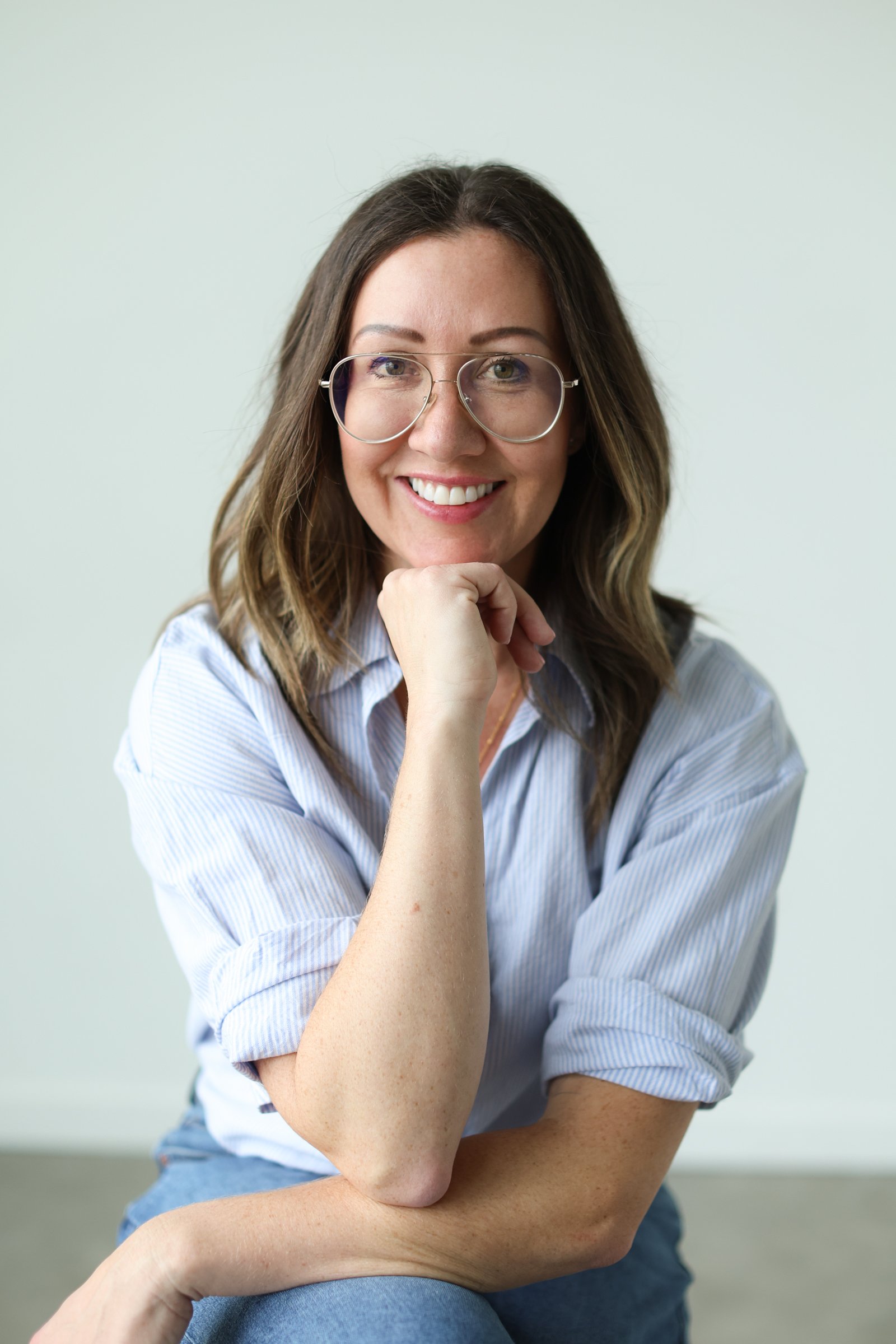A woman with shoulder-length brown hair, wearing glasses and a light blue striped shirt, smiling with her hand resting on her chin, sitting on a chair against a plain light-green background.