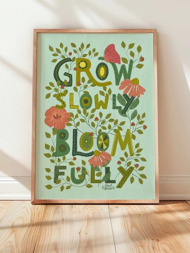 A vibrant art print with the words "Grow Slowly Bloom Fully" as large decorated letters woven together with leaves, pink wildflowers, a butterfly and tiny strawberries.