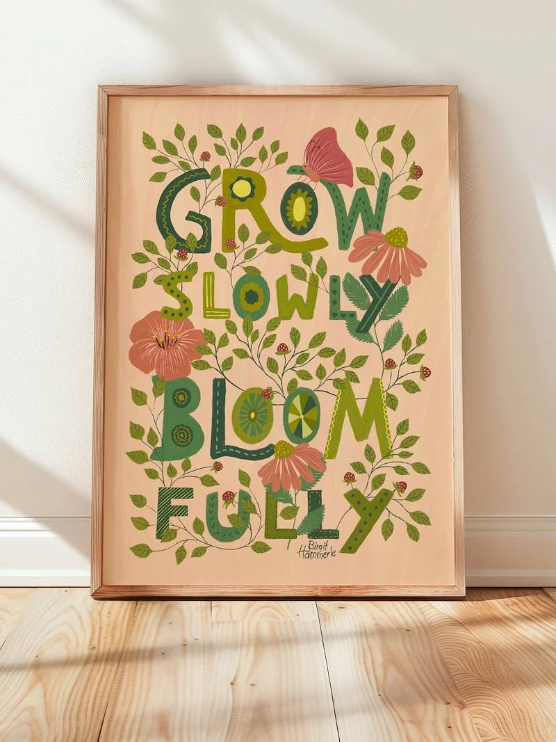 A vibrant art print with the words "Grow Slowly Bloom Fully" as large decorated letters woven together with leaves, pink wildflowers, a butterfly and tiny strawberries.