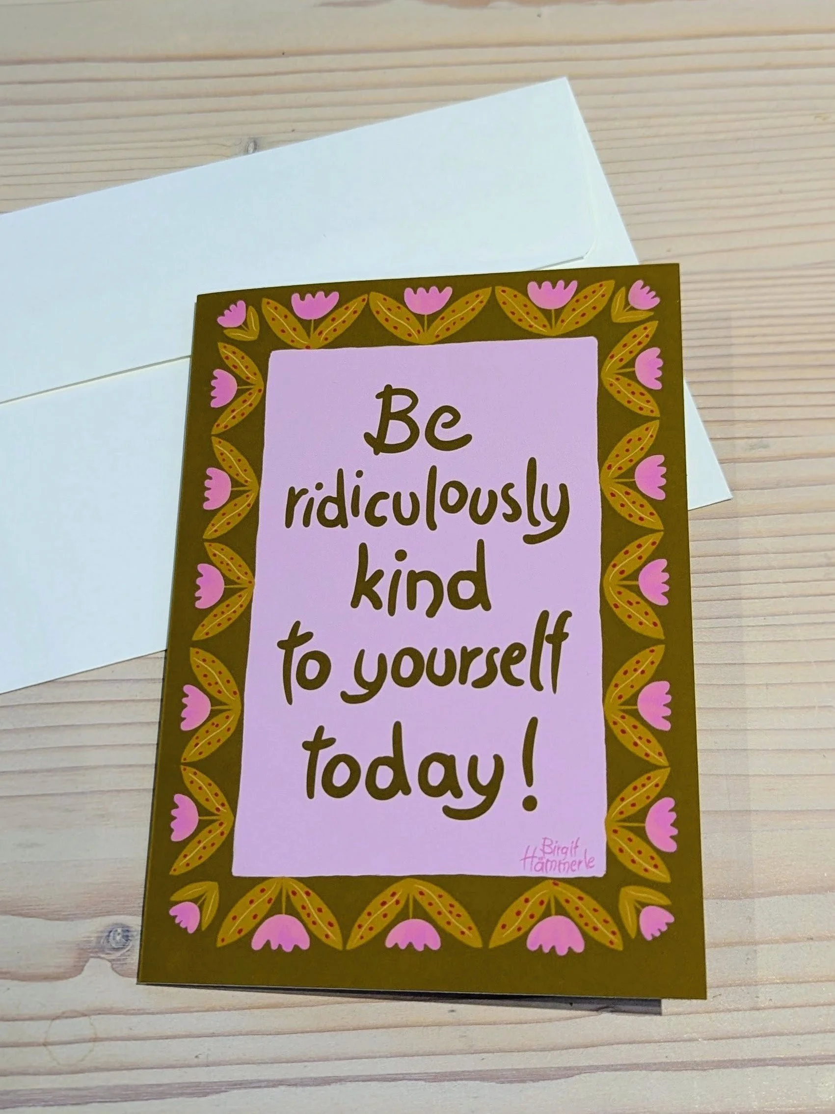 A greeting card on a wooden surface with a pink-and-green floral border. The card features handwritten text that reads, "Be ridiculously kind to yourself today!"