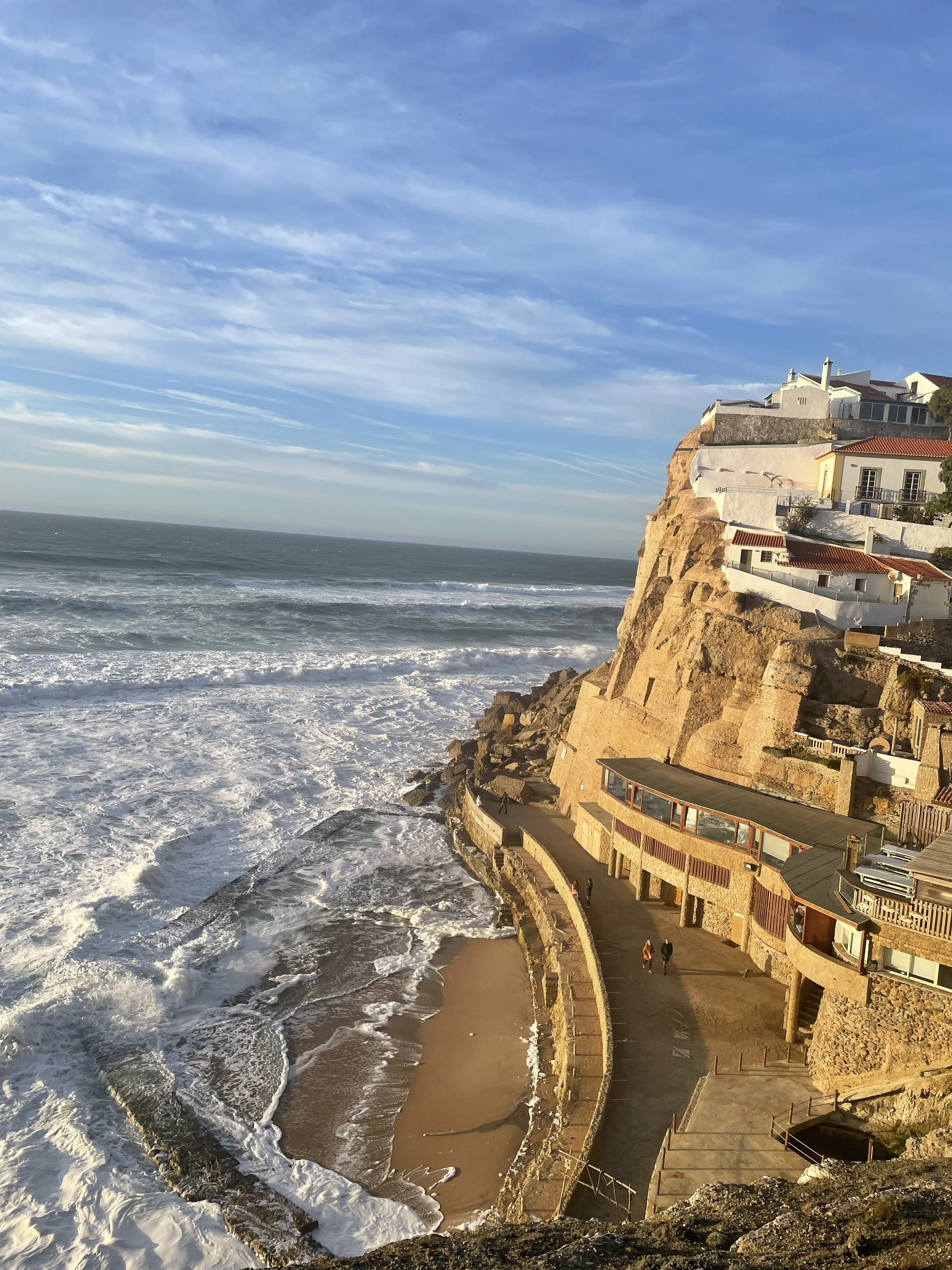 Acantilado junto al océano con construcciones en la cima, una piscina natural y un camino junto al agua. Cielo azul con nubes. Todo esto en Portugal.