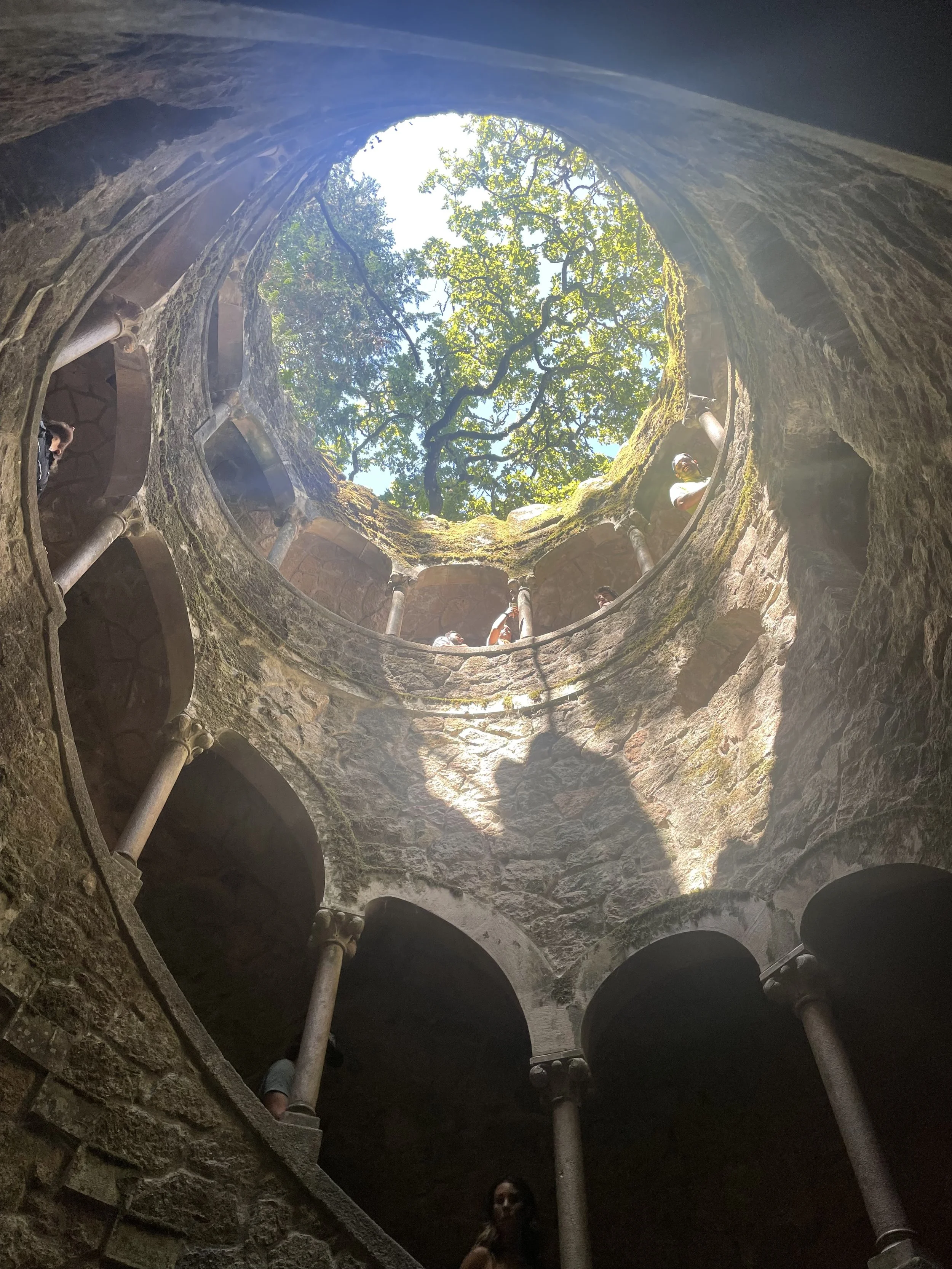 Vista desde el interior de un pozo subterráneo en espiral con columnas de piedra y vegetación al fondo en Portugal.