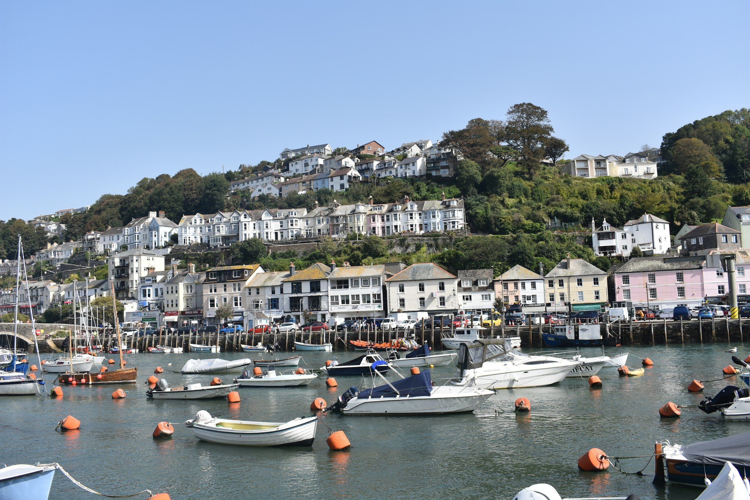 Looking out across Looe harbour with boasts moored on the Looe river and homes nestled into the hillside on a sunny day.