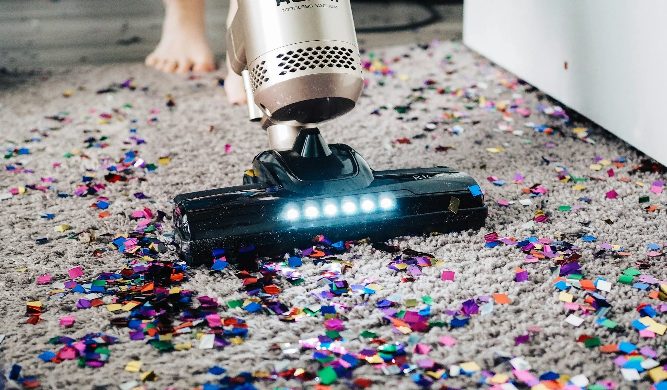 A vacuum cleaner picking up colourful confetti from a carpet, illustrating the chaos of busy households and the need for professional help.