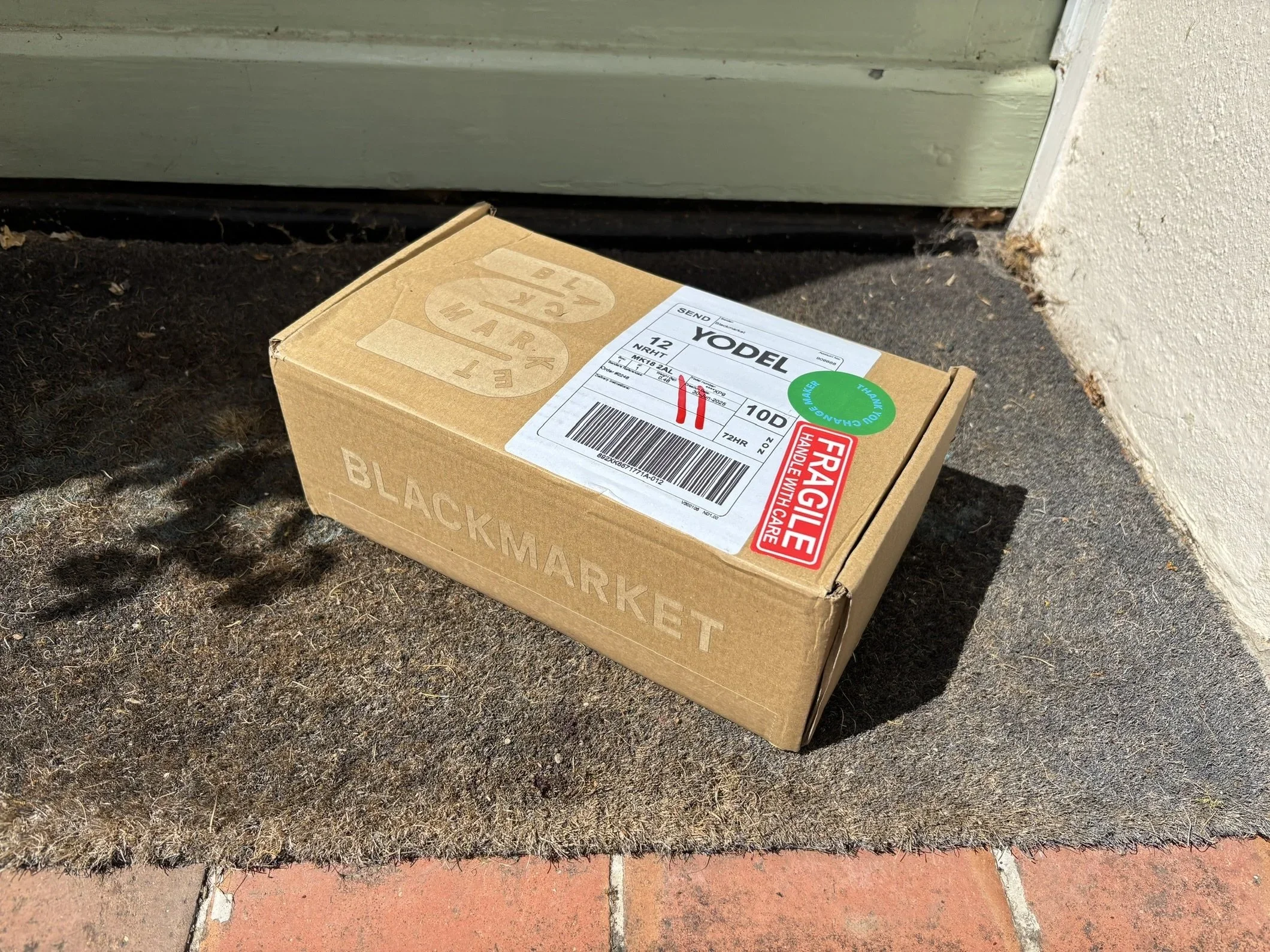 A cardboard box labeled 'Black Market' is on a doormat outside a door on a concrete porch.