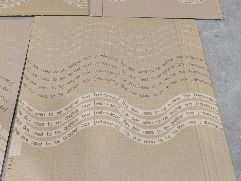 Multiple light brown cardboard boxes with printed text, some stacked and others lying flat on a concrete surface. The boxes have repetitive text, including 'This box used to be another box' and '[rebox.eco]', indicating eco-friendly packing materials.