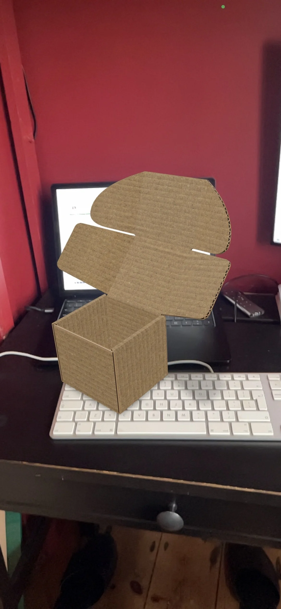 A cardboard box with an open lid, placed on a computer keyboard on a desk. The background shows a monitor, a laptop, and a remote control against a red wall.