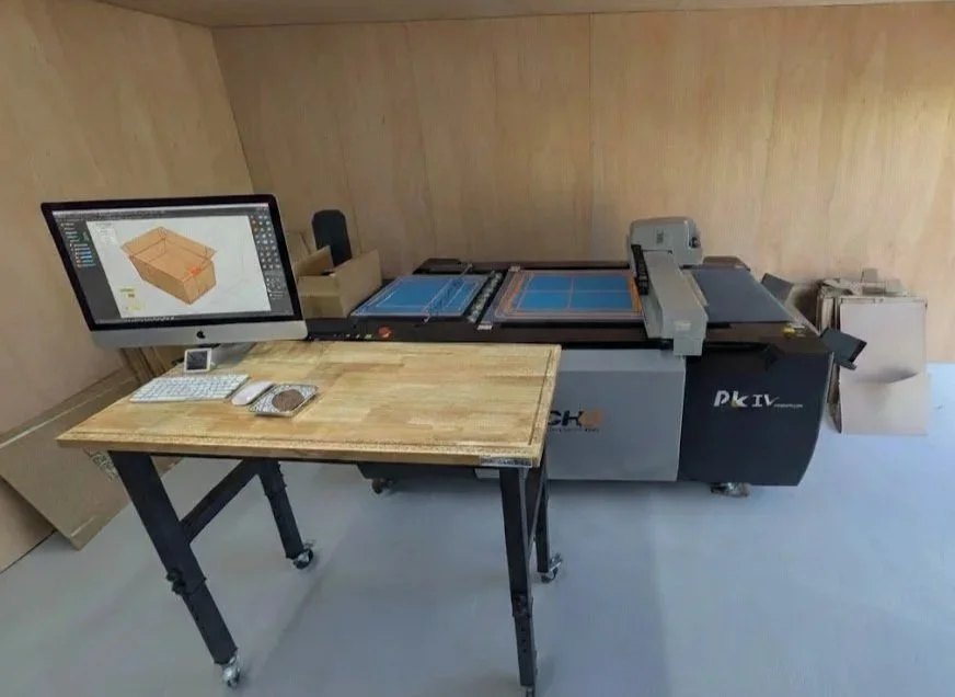 Laser cutting machine in a workshop with a computer display showing a 3D model of a wooden box.