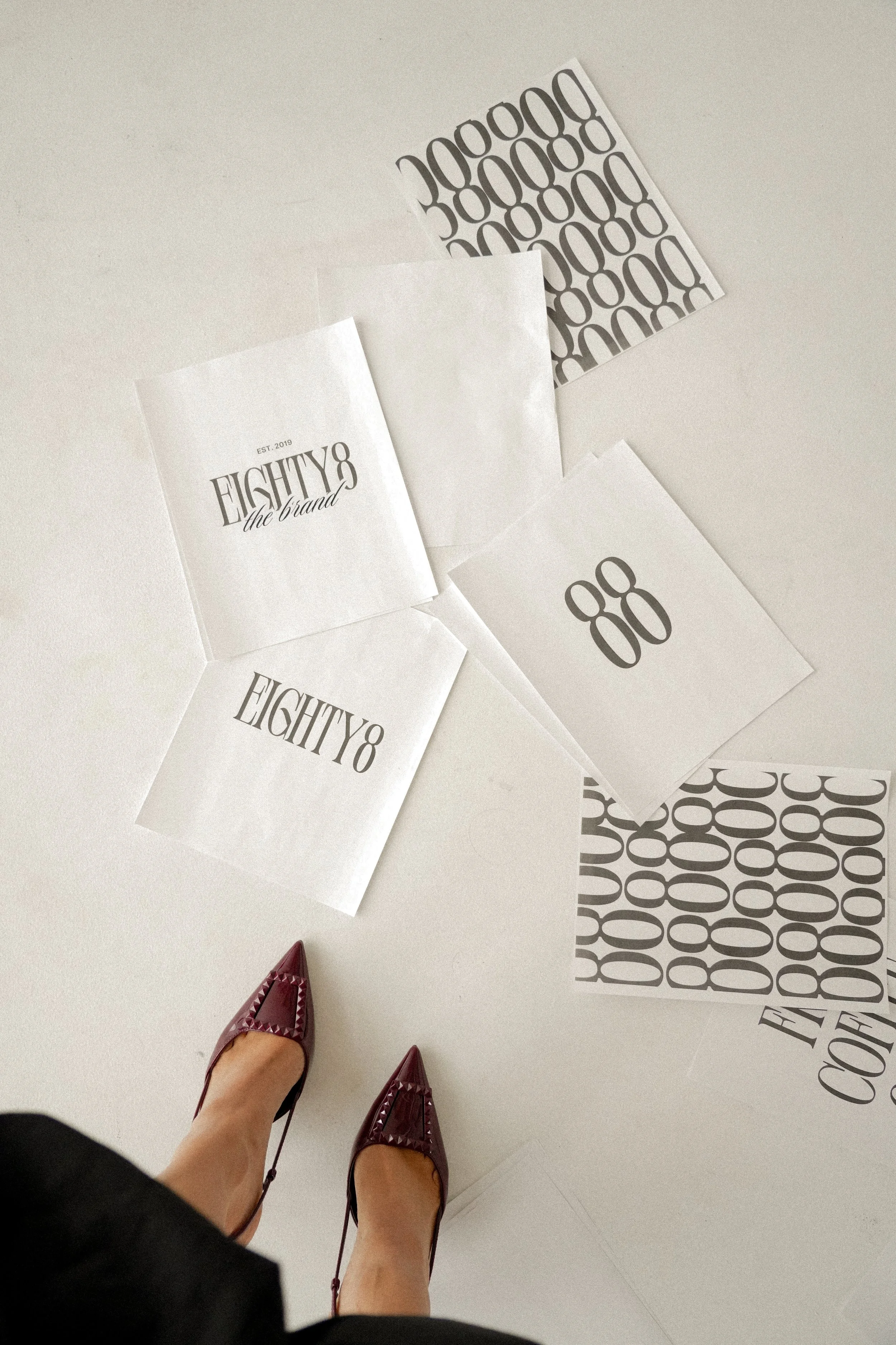 Photo of a person standing on a white floor surrounded by sheets of paper with black text and graphics. The person's feet are visible, wearing burgundy pointed-toe shoes with decorative stitching, and a black skirt or dress.