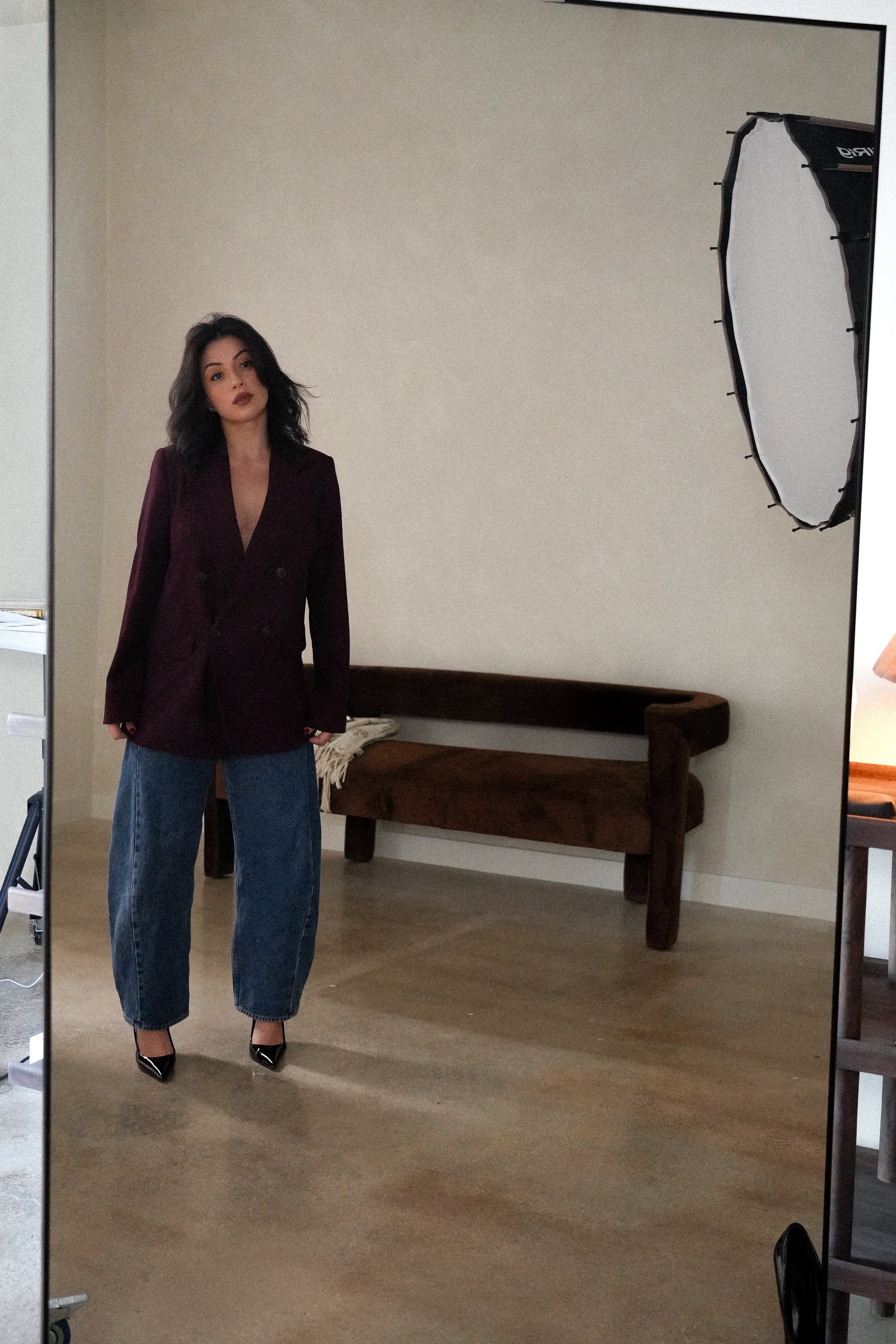 A woman taking a mirror selfie in a room with a beige wall, a wooden bench, and a photography softbox.