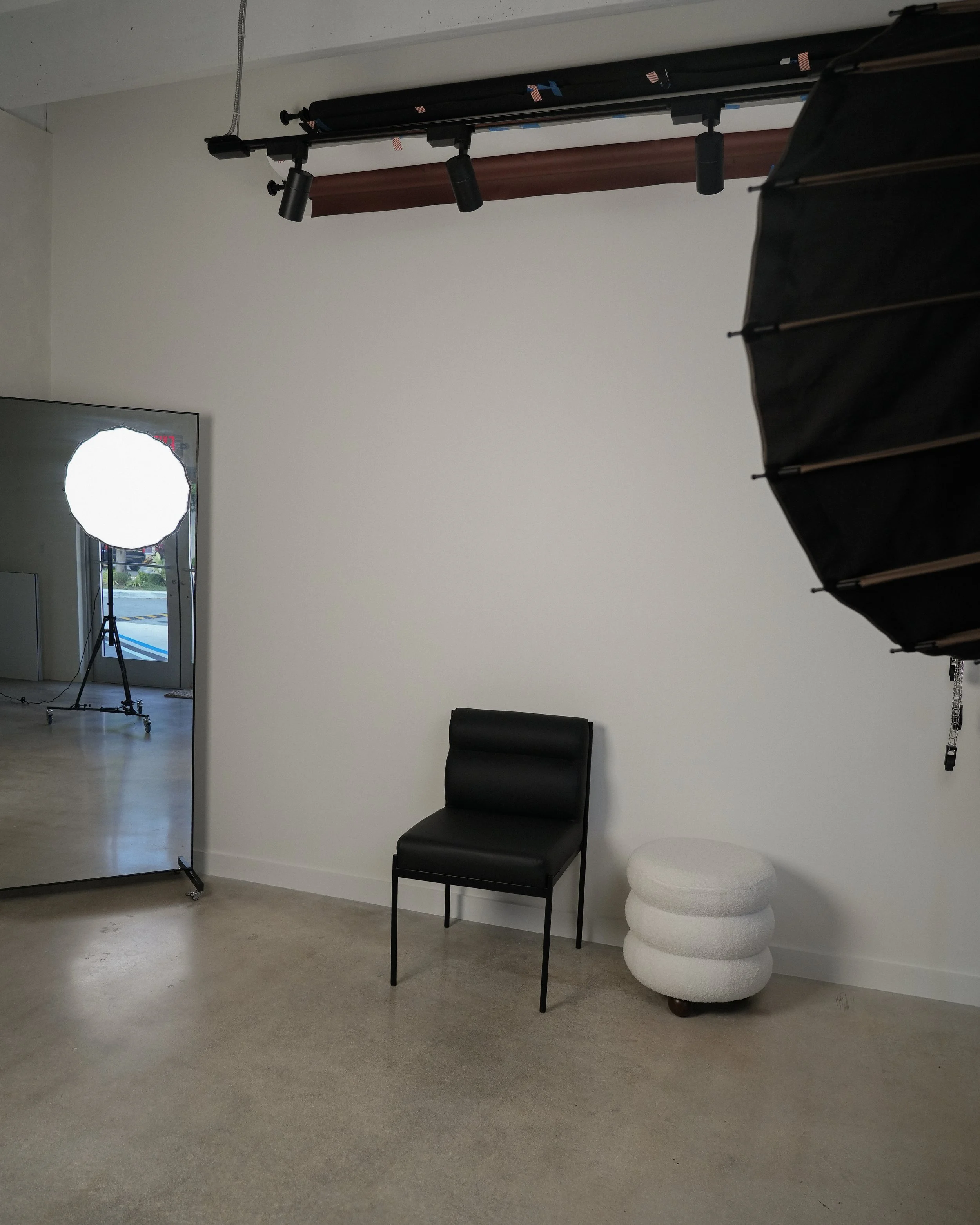 Studio setup with a black chair, white textured stool, photography lighting equipment, and a large mirror in a room with plain white walls.
