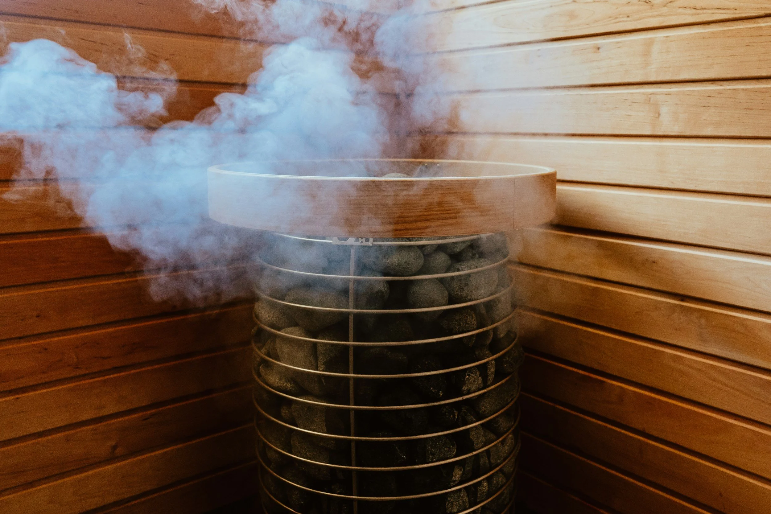 Steam rising from a sauna heater filled with dark stones, enclosed in a wire cage, set against wooden wall panels.
