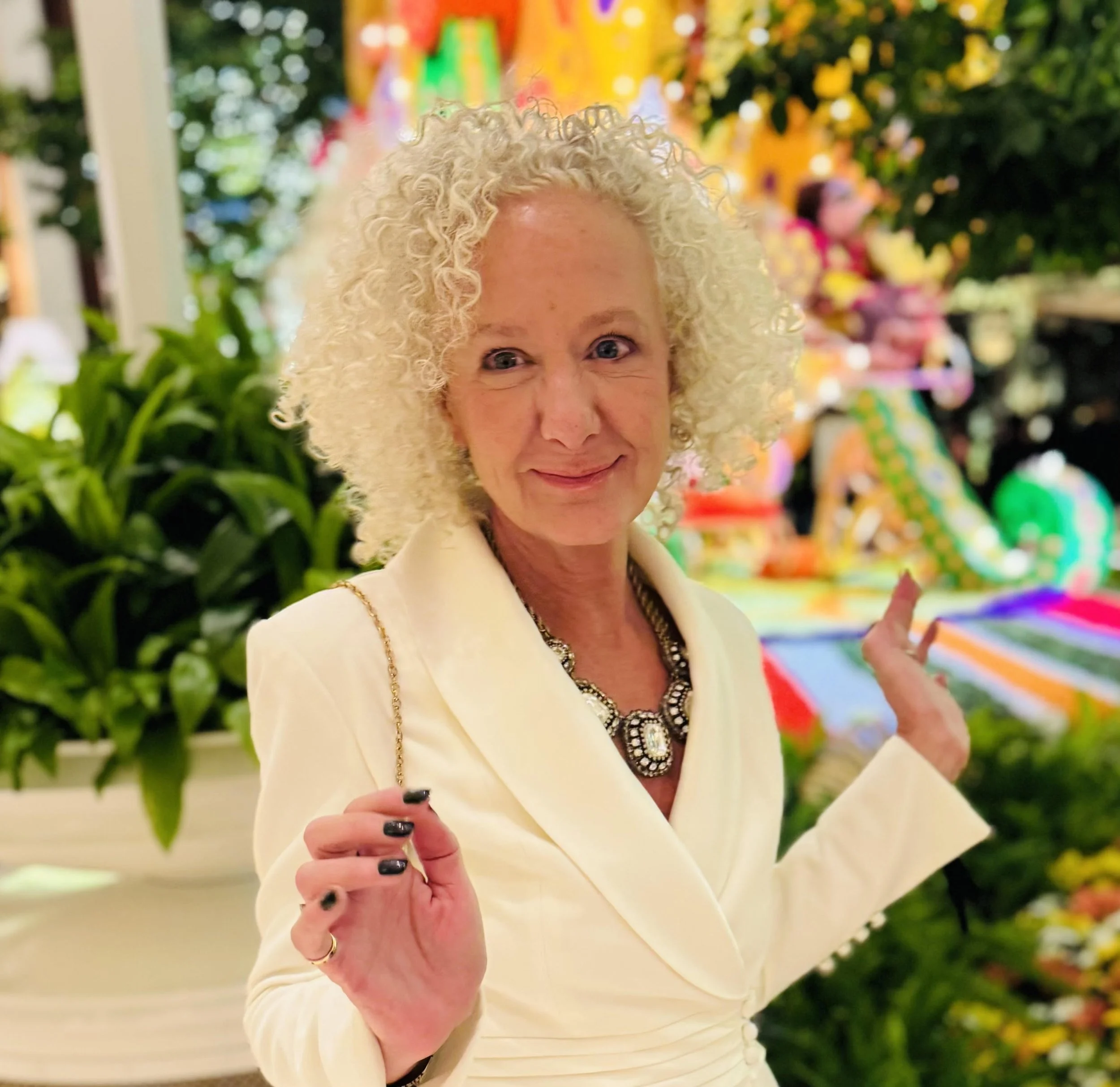 A woman with curly blonde hair wearing a cream-colored blazer and a large necklace, standing in front of colorful decorations and plants, smiling and gesturing with her hands.