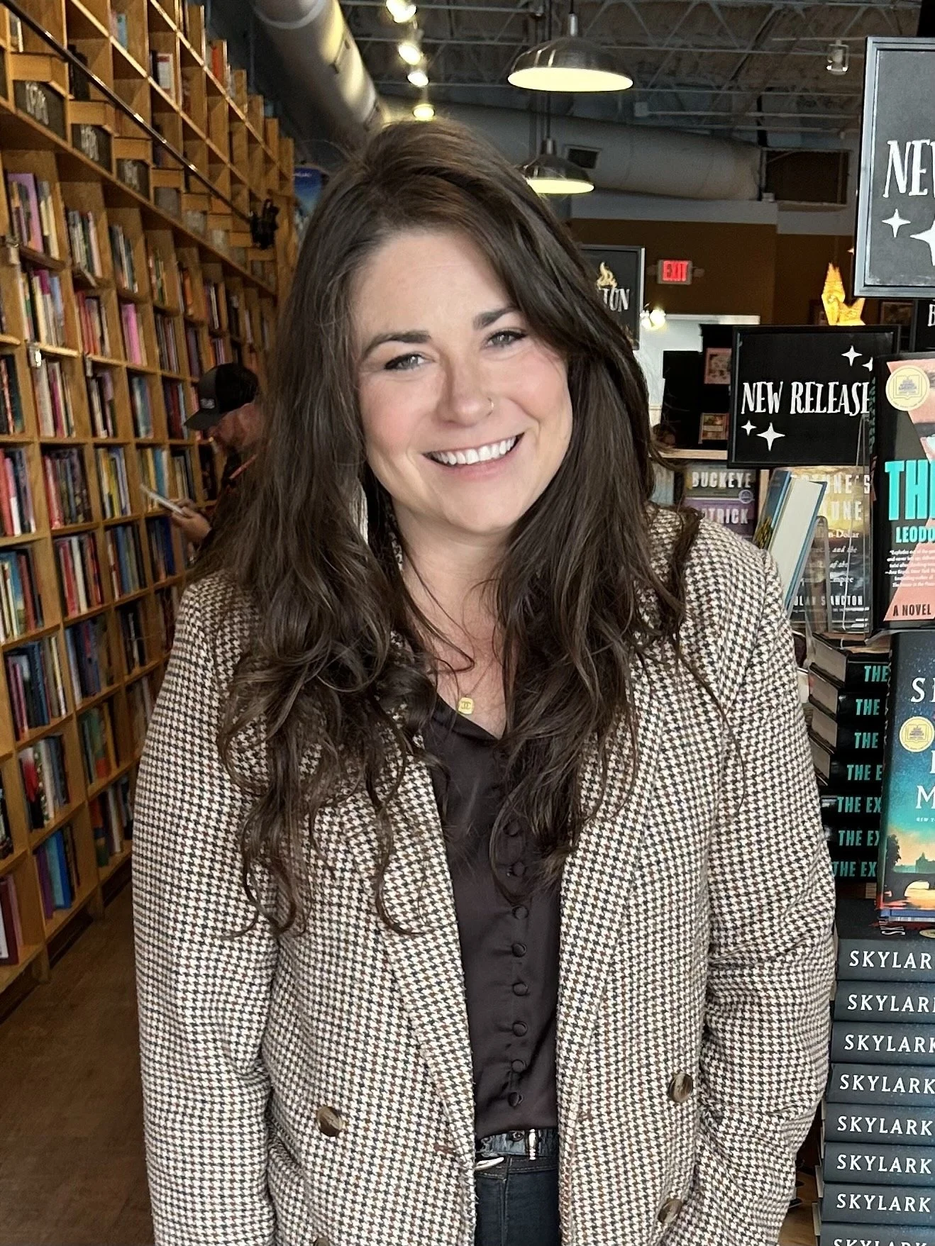 A young woman with long dark hair smiling inside a bookstore, standing in front of shelves filled with books.